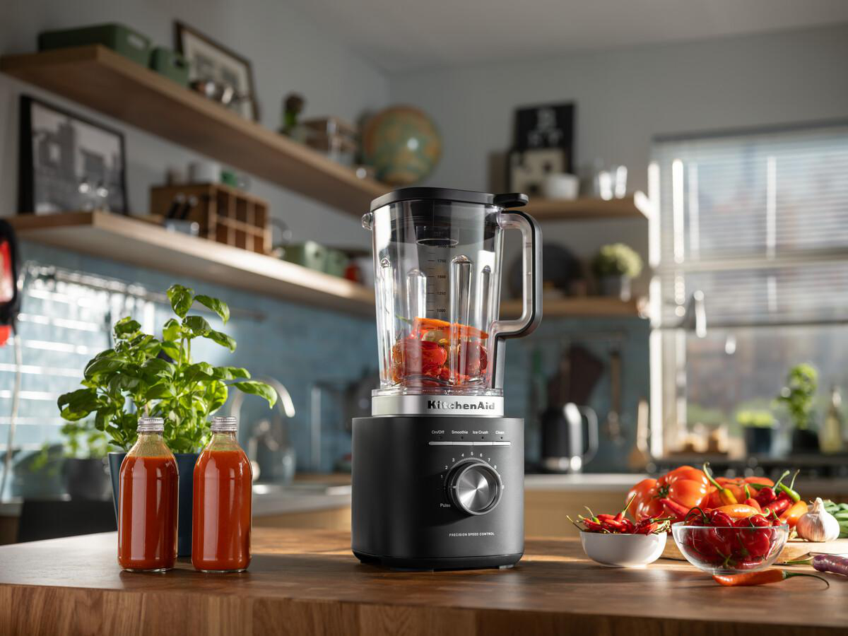 Ein schwarzer KitchenAid-Mixer mit Gemüse darin, neben zwei Soßenflaschen, Tomaten, Paprika und einer Pflanze auf einer Holztheke.