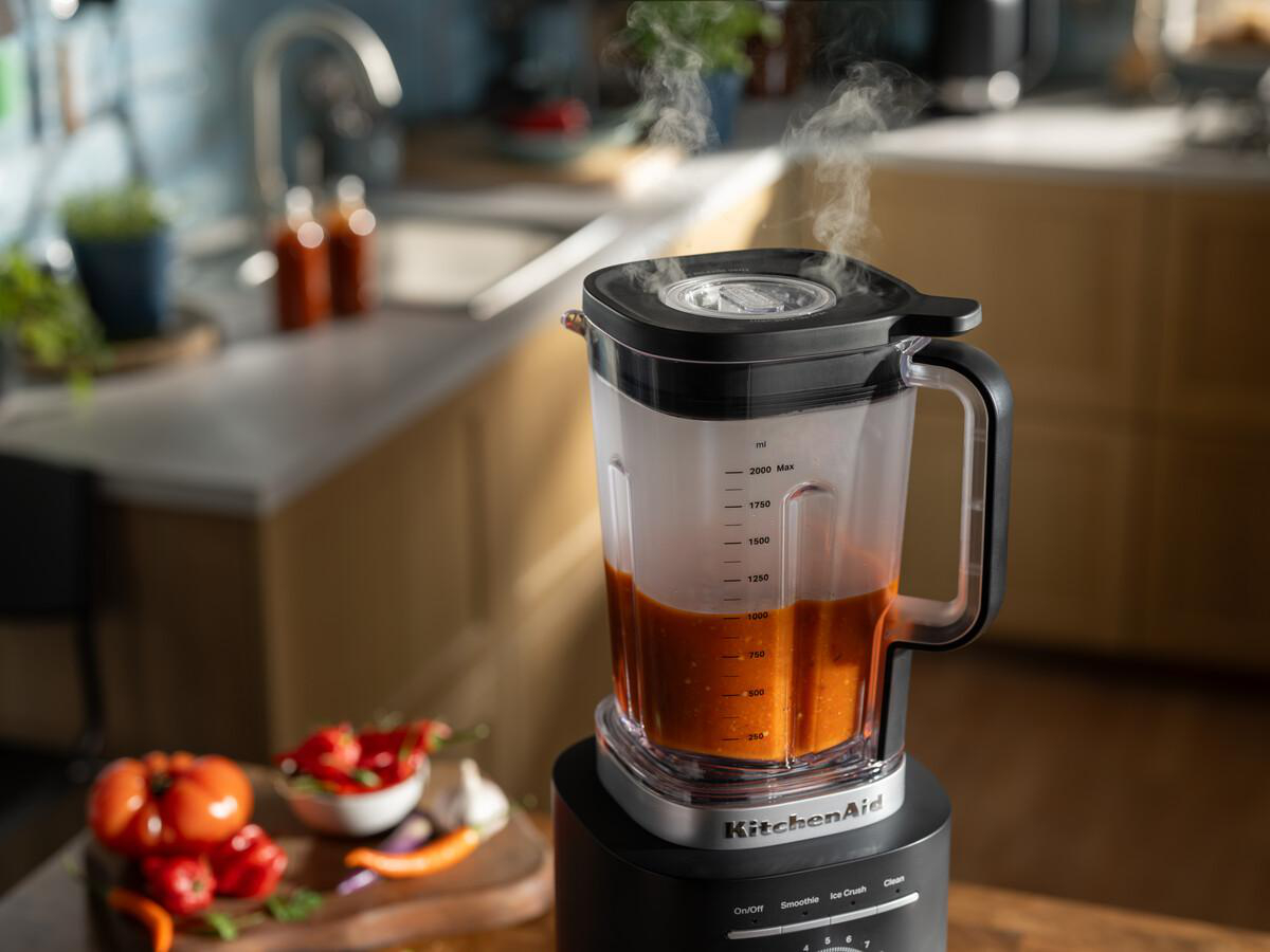 Ein Mixer mit Tomatensauce steht auf einer Küchentheke. Dampf steigt aus dem Mixer auf. Tomaten und Paprika liegen auf einem Holzbrett.
