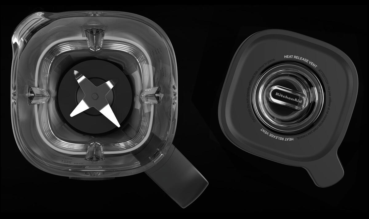 Ein klarer Mixbehälter mit Klinge und Deckel. Der Deckel hat ein KitchenAid-Logo und eine Wärmeableitung. Der Hintergrund ist dunkel.