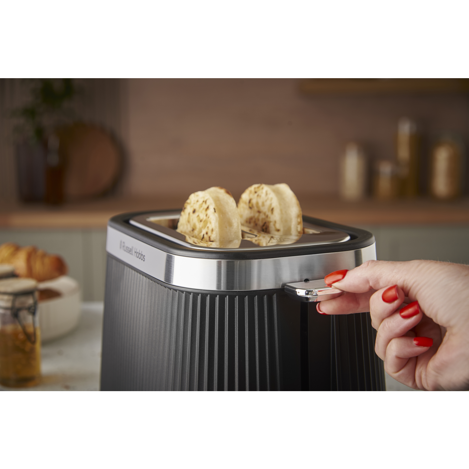 A black toaster with two slices of toasted bread is shown. A hand with red nails is pushing the lever down.