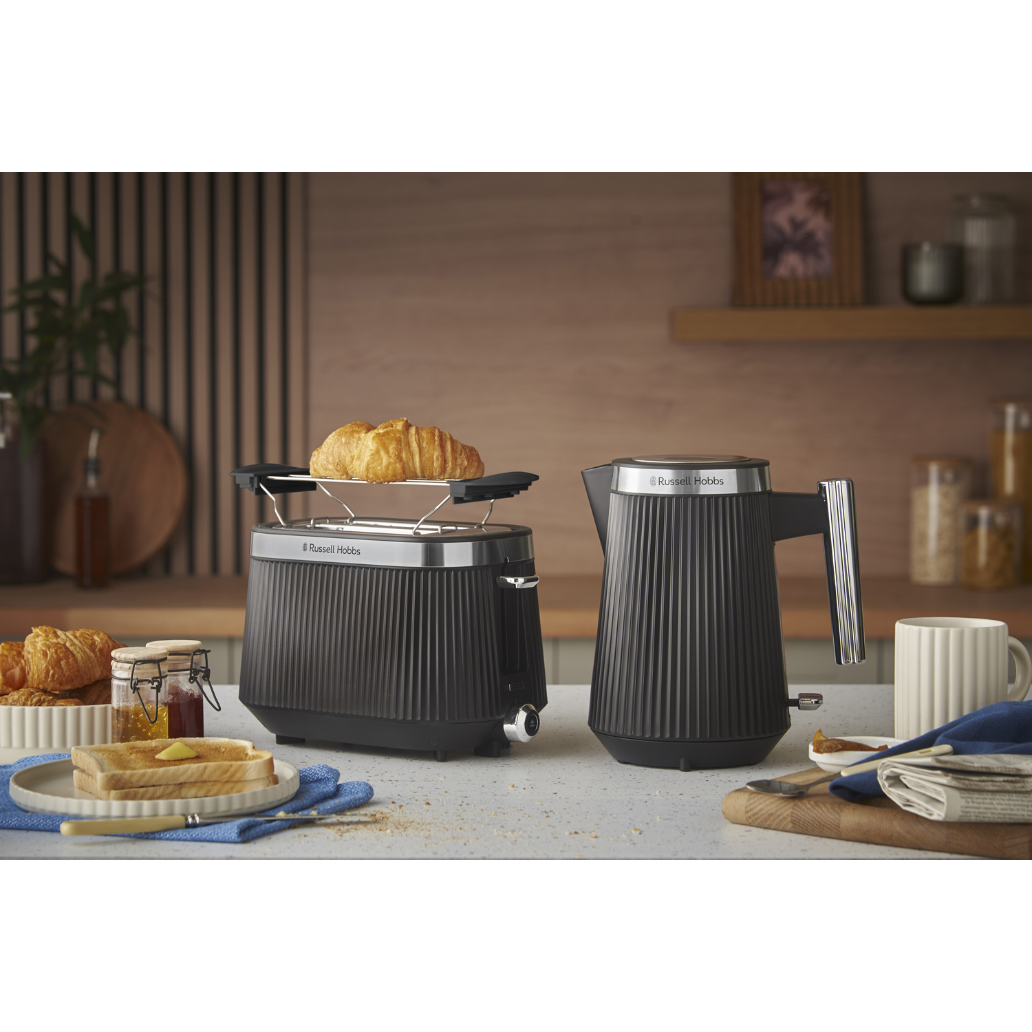 A kitchen scene with toaster, kettle, toast, and croissants. A jar of jam and a mug are also on the counter.
