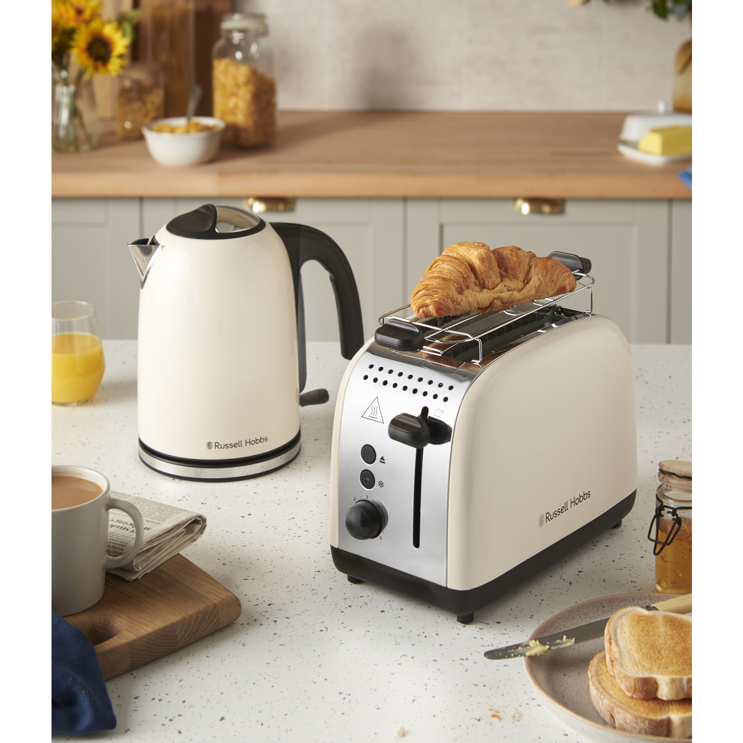A toaster with a croissant on top, a kettle, coffee, toast, and jam are on the counter, with a wooden cutting board.