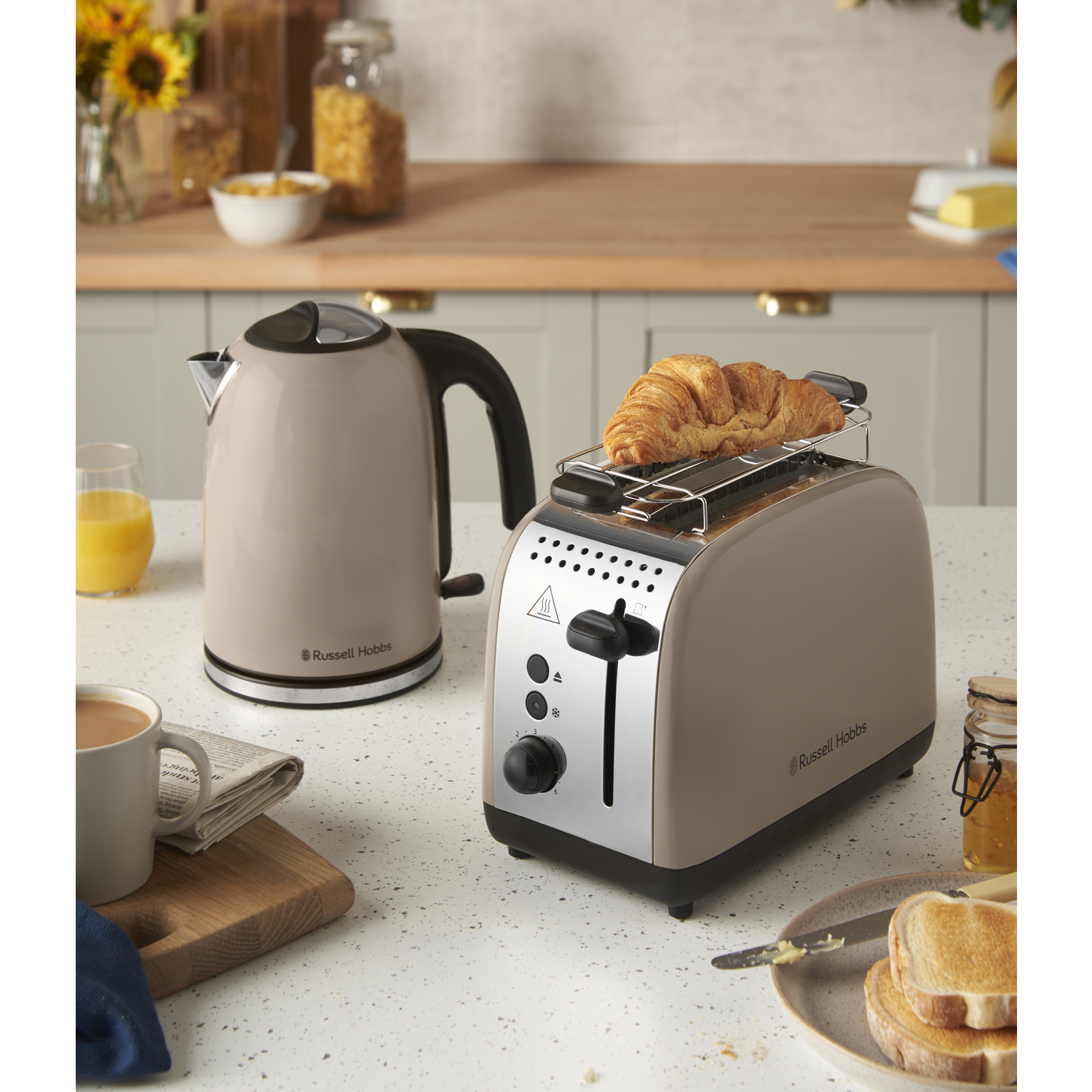 A kitchen scene with a toaster holding a croissant. Beside it, a kettle, coffee, and toast with butter are present.