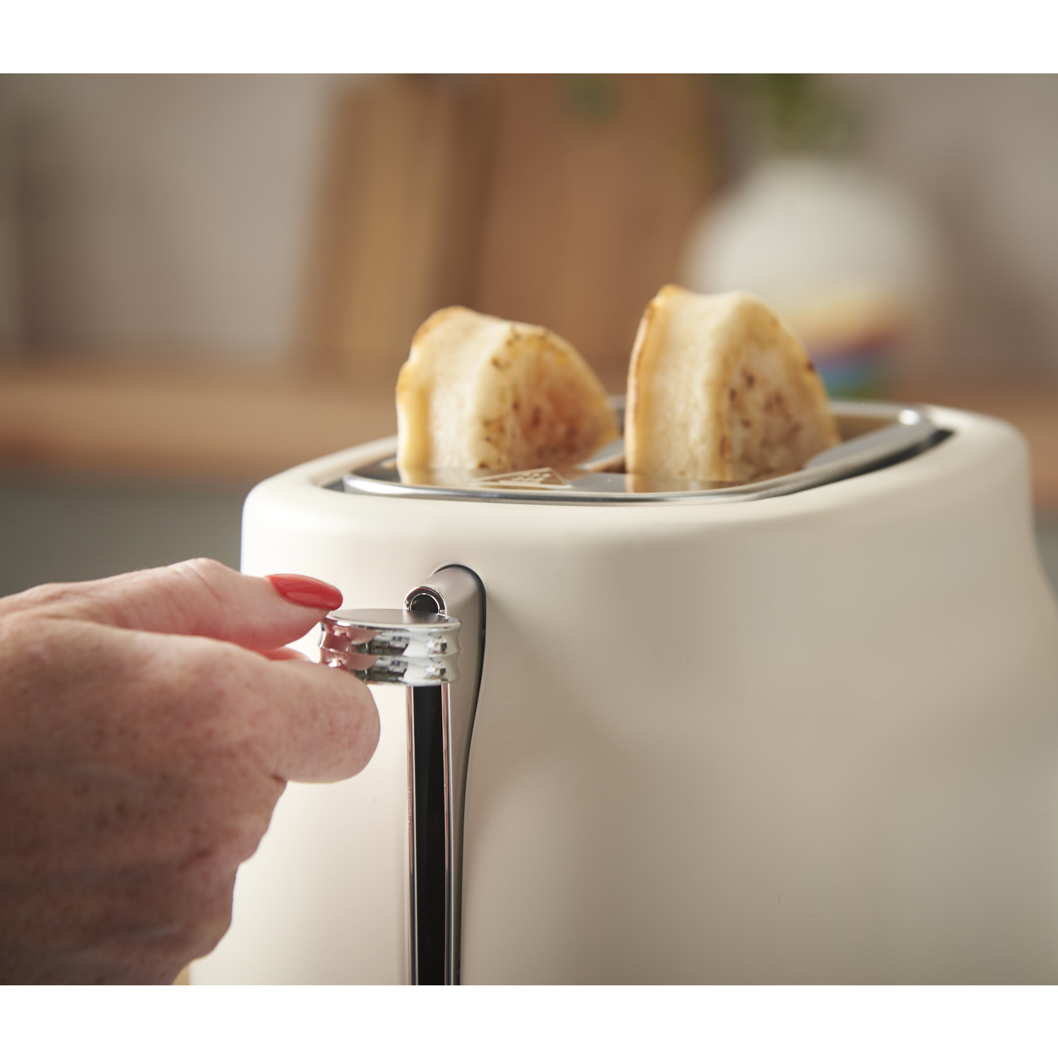 A hand with red nail polish adjusts a cream-colored toaster. Two pieces of toast are inside. Background has a blurred vase.