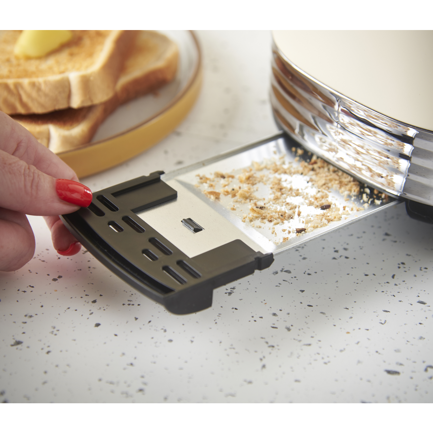 A hand with red nail polish removes a crumb tray from a toaster. Toasted bread with butter is on a plate in the background.