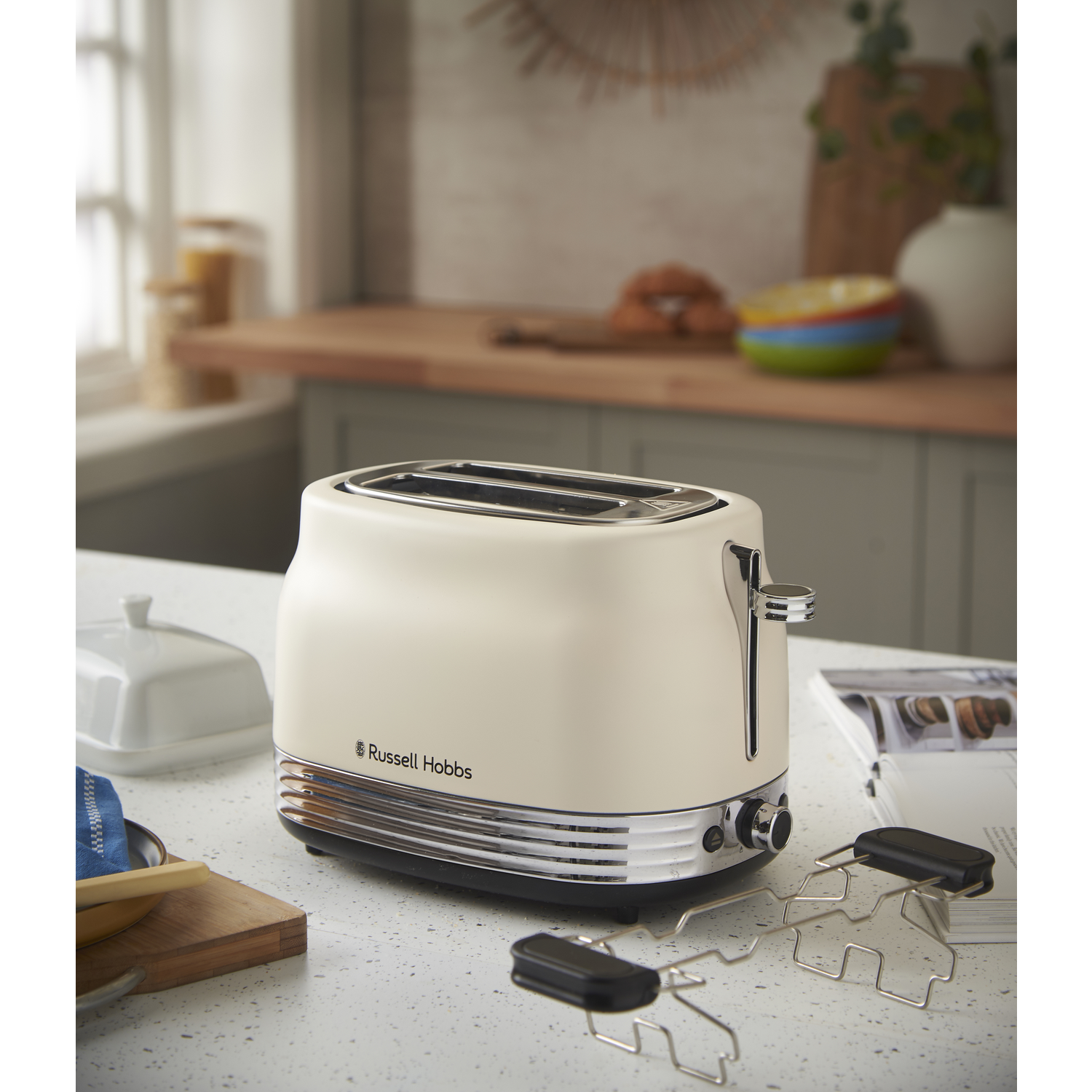 A cream-colored Russell Hobbs toaster sits on a countertop. Accessories and other kitchen items are visible in the background.
