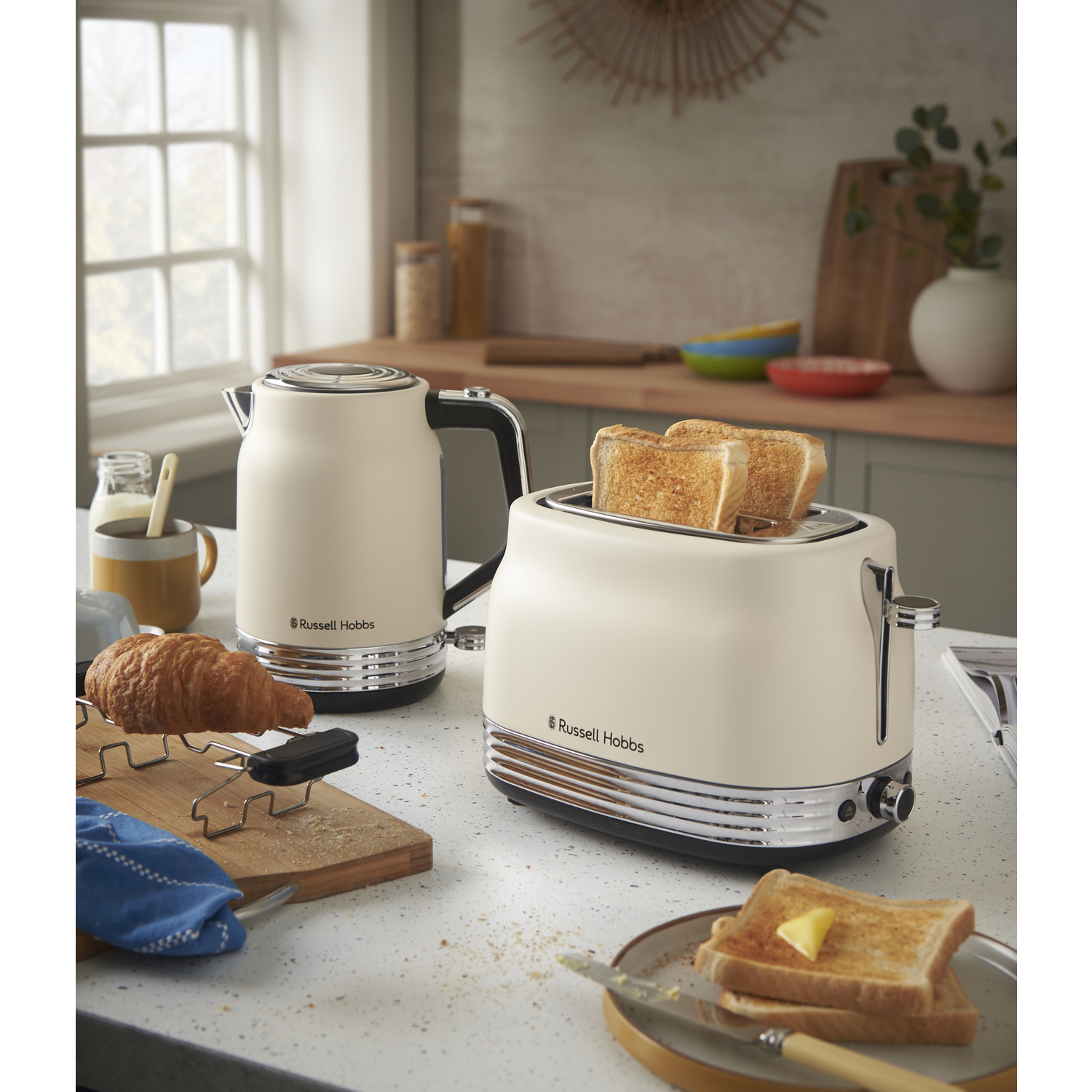 A kitchen scene shows a toaster with toast, a kettle, and a croissant on a cutting board. Sliced toast with butter is on a plate.