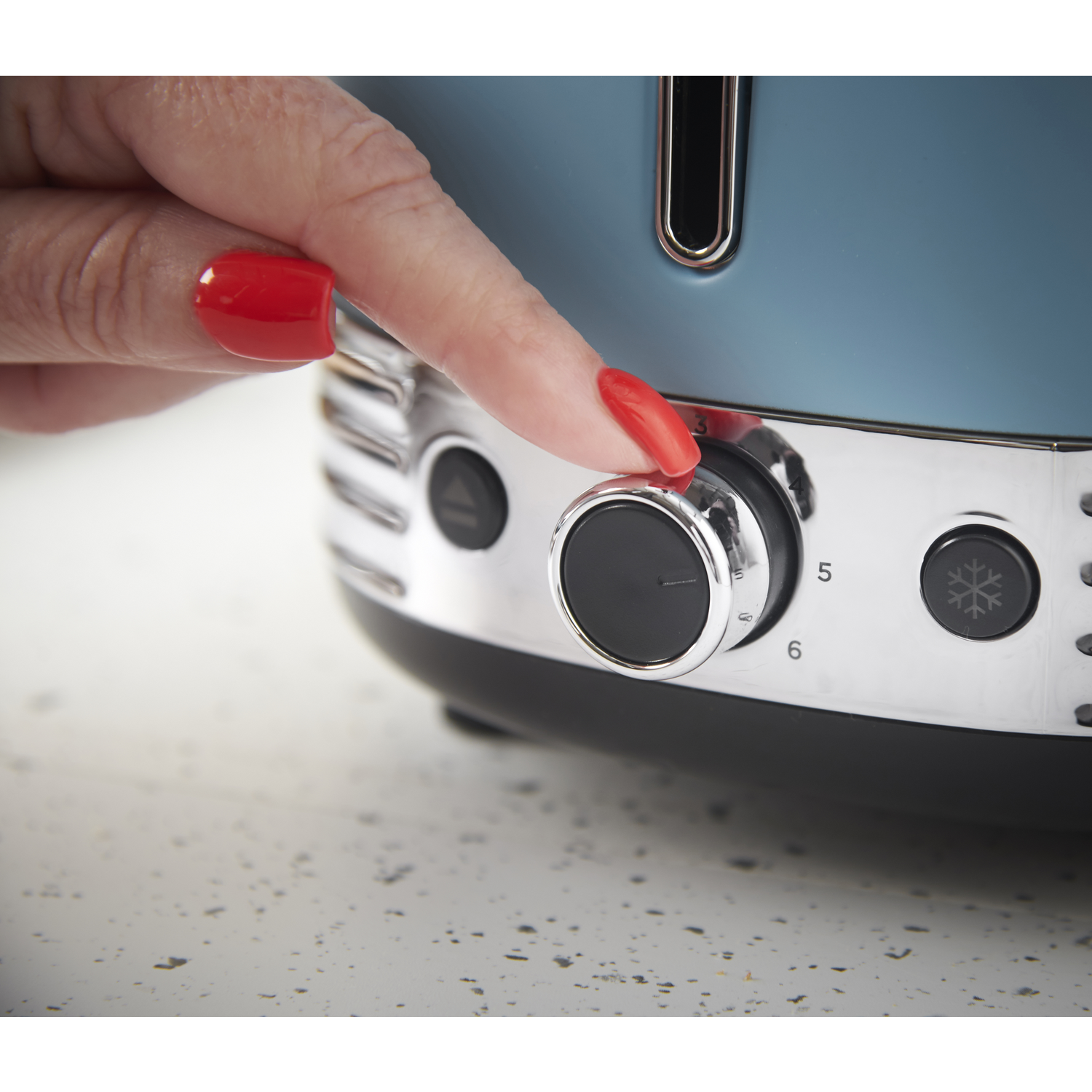 A hand with red nails adjusts a toaster dial. The toaster is blue and chrome, set on a white speckled surface.