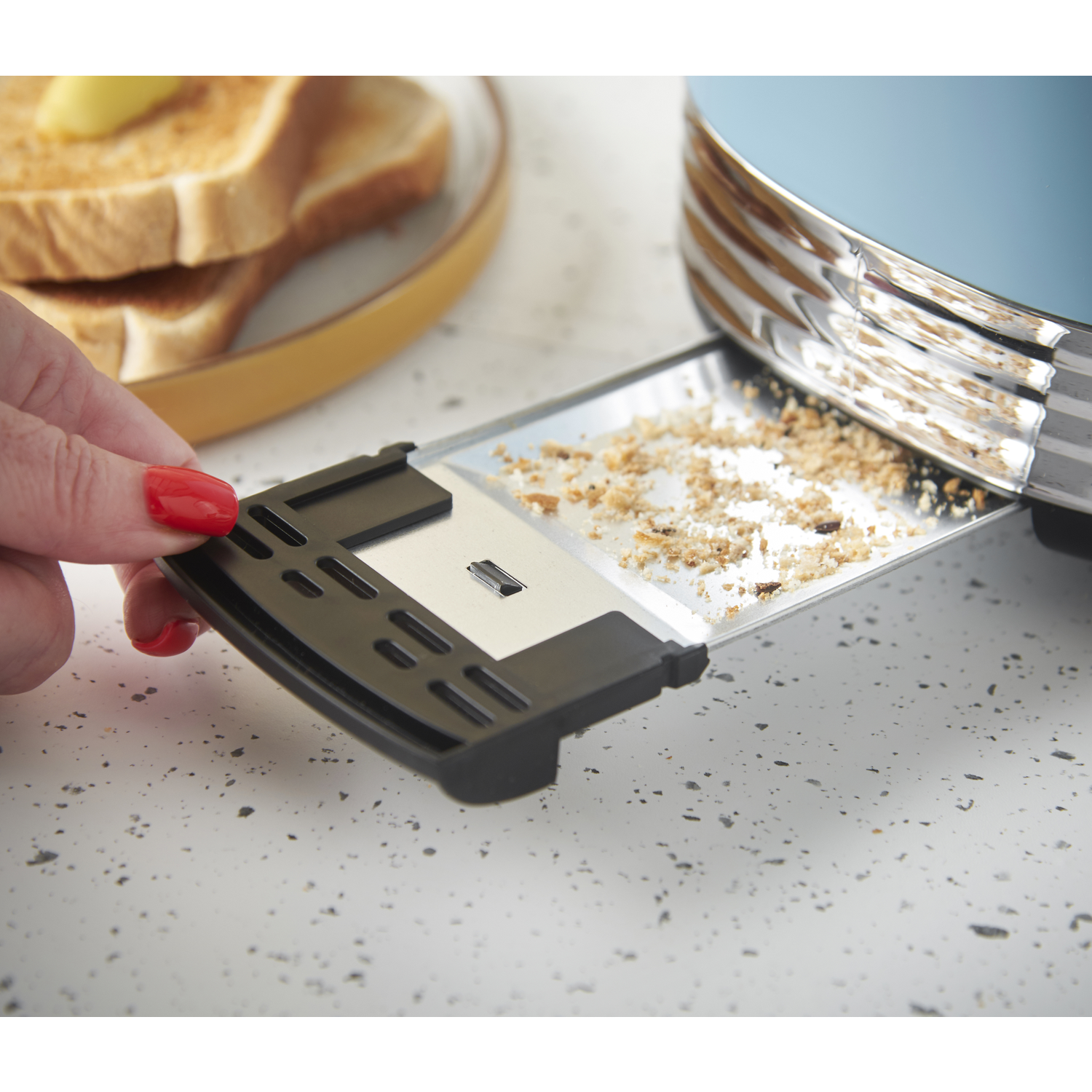A hand with red nail polish pulls out a crumb tray from a toaster. Toast with butter sits on a plate.