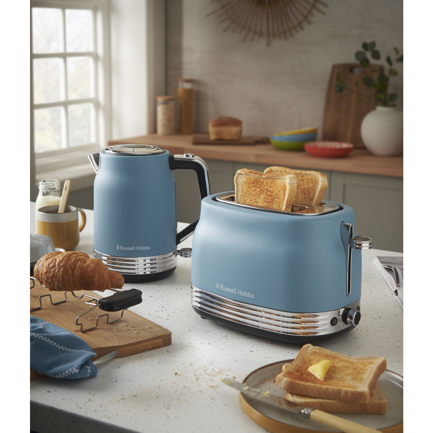 A blue kettle and toaster with toasted bread. A croissant and buttered toast are on the counter, kitchen background.