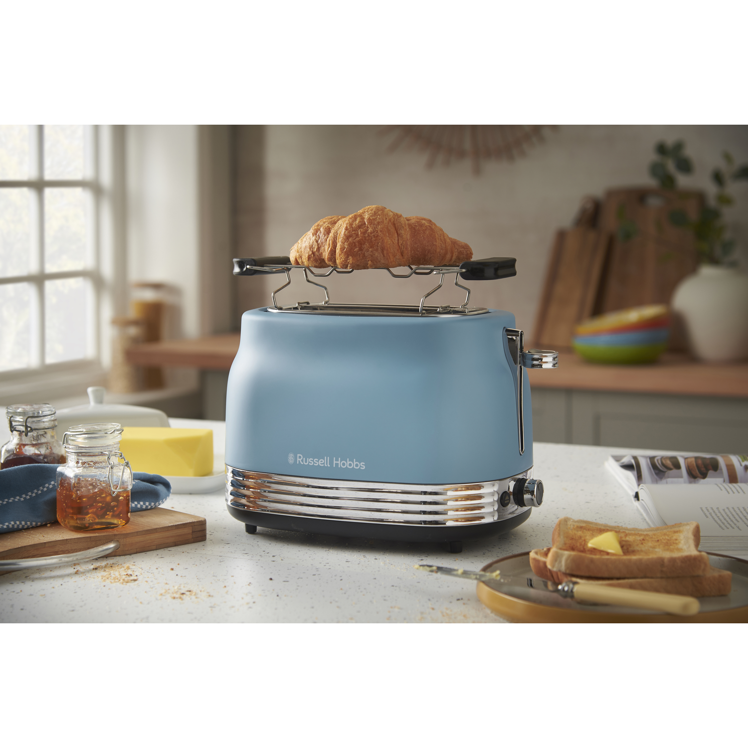 A blue toaster with a croissant on top. Toast and butter are on a plate, with jam and butter on the side.