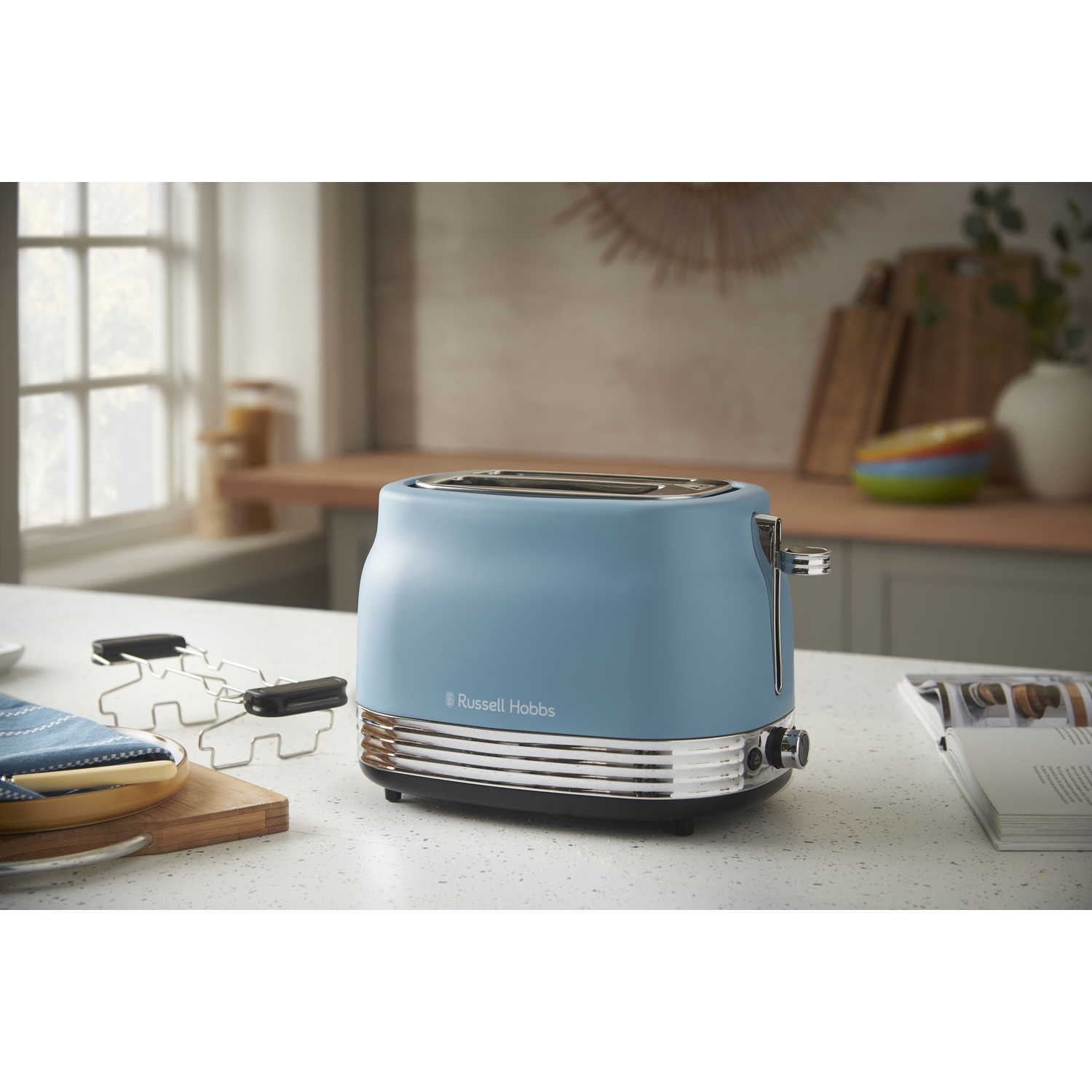 A blue Russell Hobbs toaster on a kitchen counter. A bread rack, a plate with a knife, and an open book are nearby.