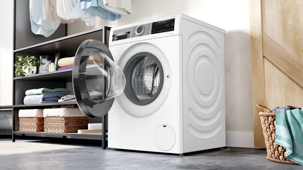 Biała pralka z otwartymi drzwiami. Półki z ręcznikami i roślinami po lewej. Kosz z ręcznikami po prawej.