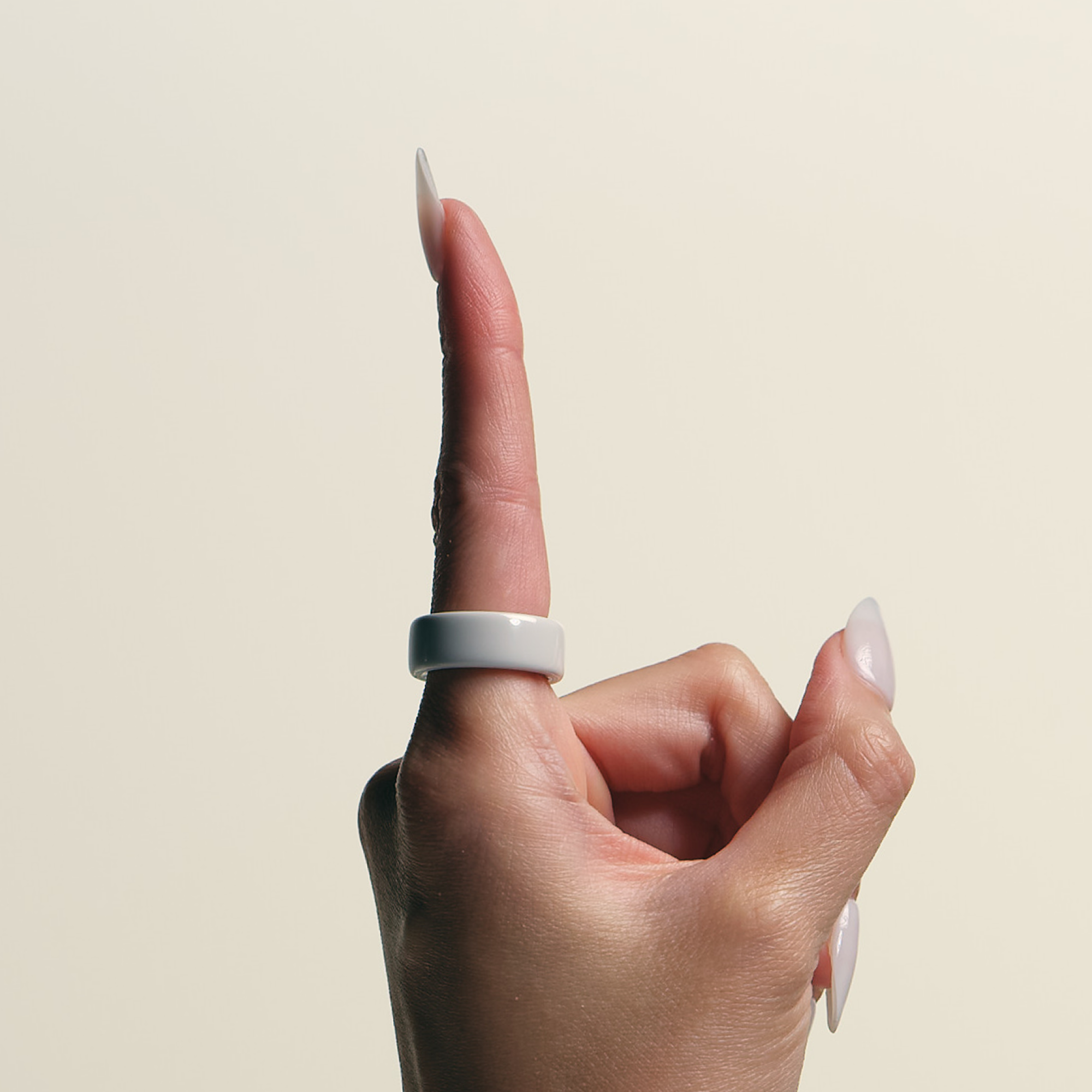 Una mano con unghie lunghe, con un anello bianco, indica verso l'alto. Lo sfondo è beige chiaro uniforme.