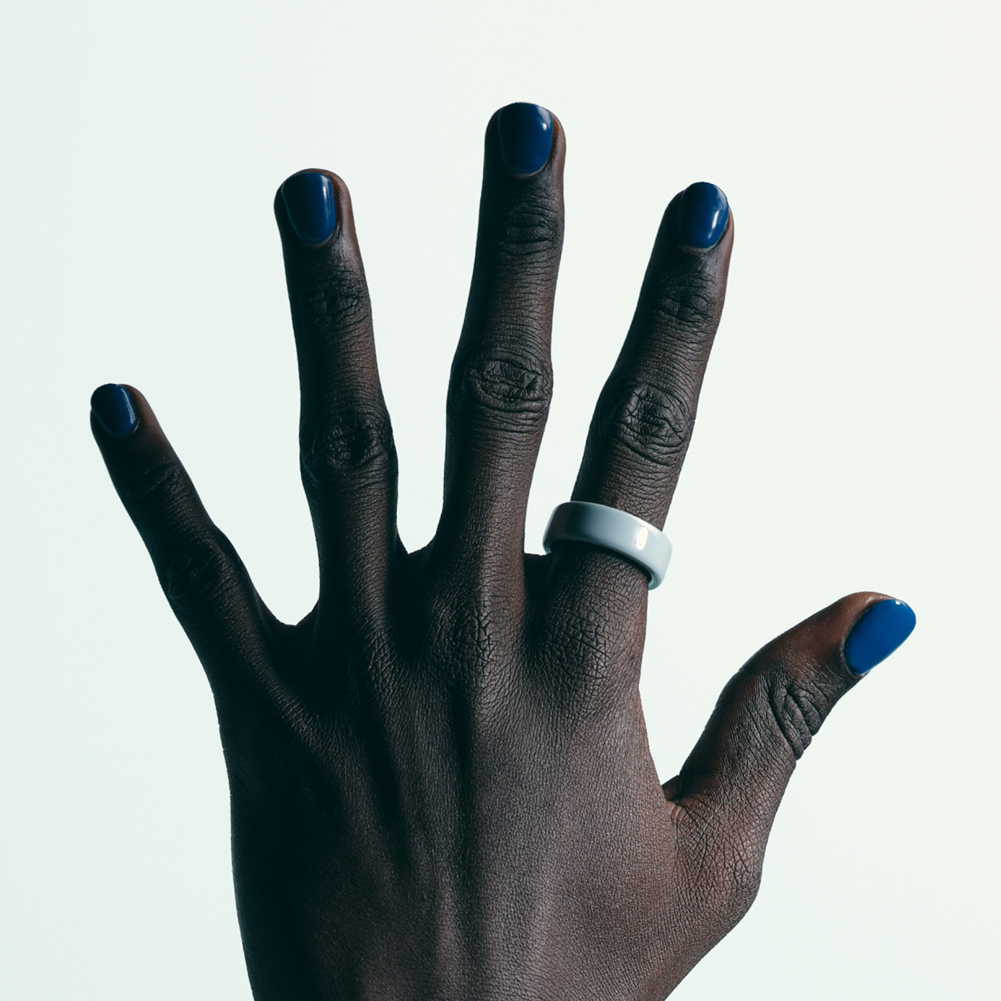 Una mano scura con unghie blu e un anello azzurro su sfondo bianco, dita divaricate.