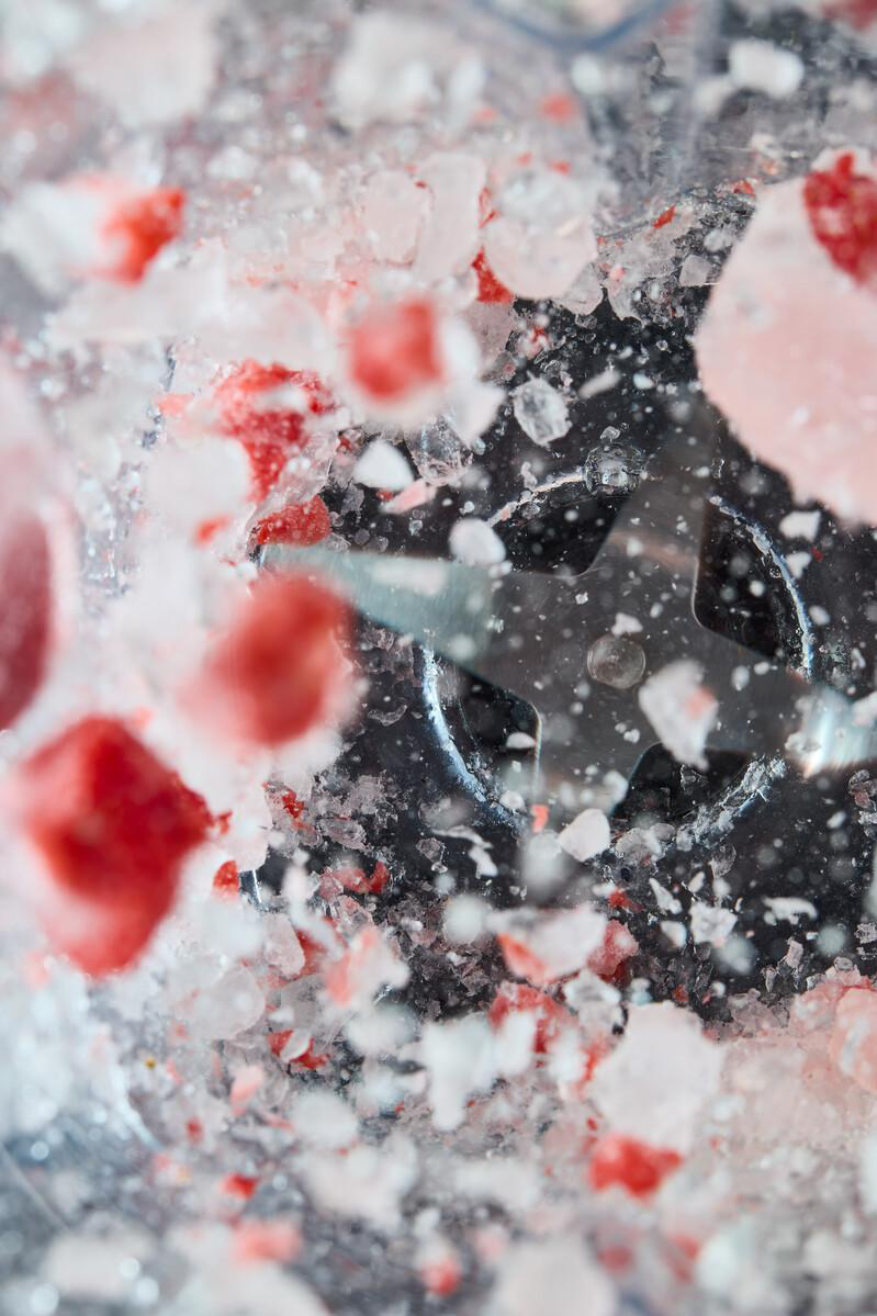 Nahaufnahme eines Mixers mit Eis und roten Fruchtstücken. Ein vierblättriges Metallteil ist sichtbar. Die Szene ist eine Mischung aus Rot, Weiß und Schwarz.
