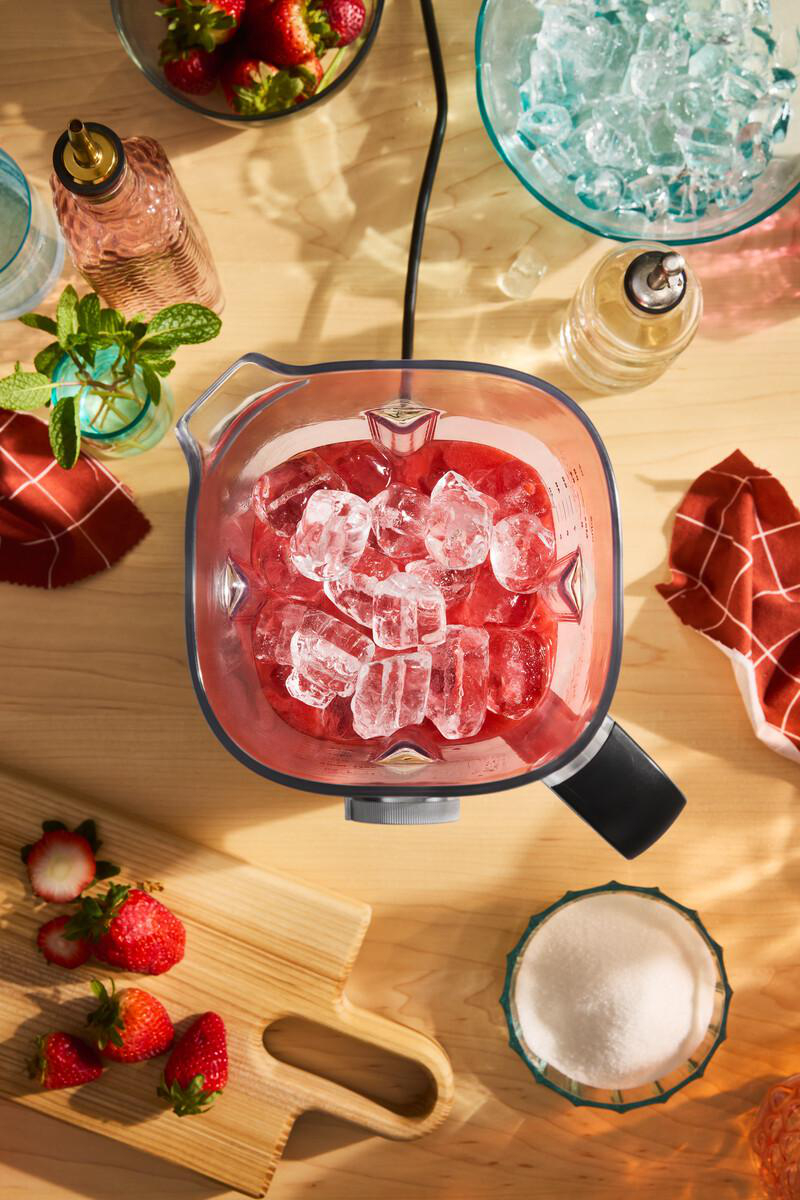 Draufsicht auf einen Mixer mit roter Flüssigkeit und Eis. Erdbeeren, Eis und Zucker liegen in der Nähe auf einem Holztisch.