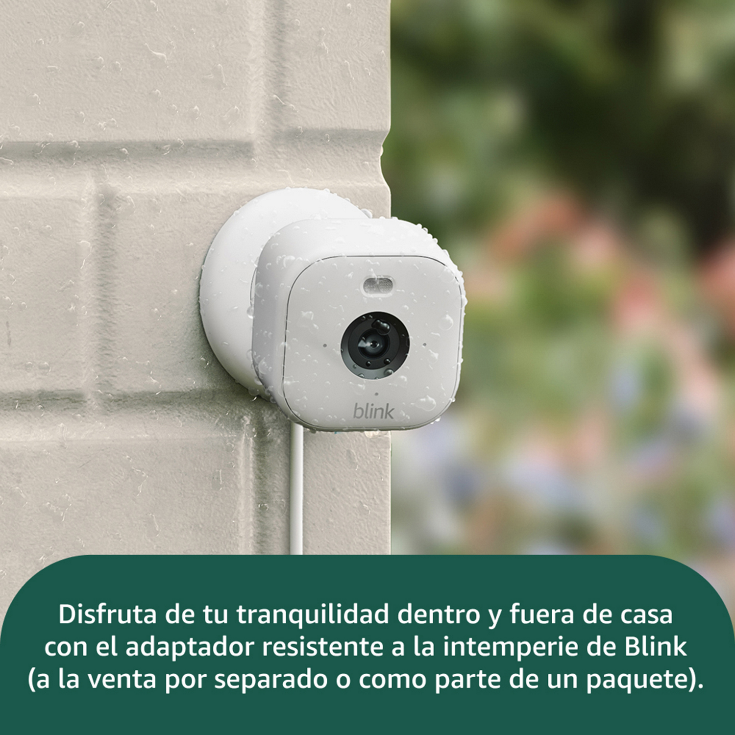 Una cámara de seguridad Blink blanca está montada en una pared de ladrillo claro. Gotas de agua son visibles en la cámara. Follaje verde borroso en el fondo.