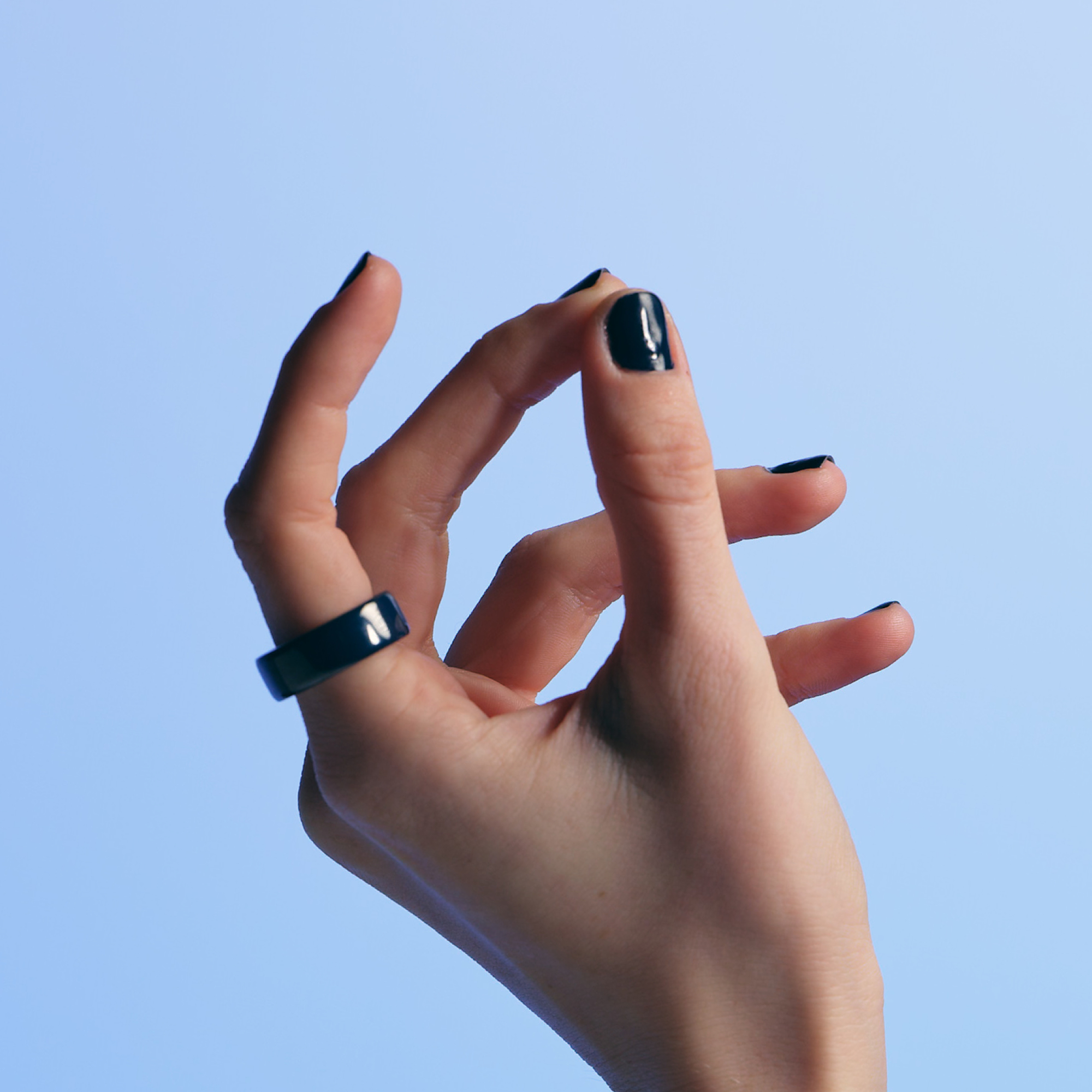 Eine Hand mit schwarzem Nagellack und einem schwarzen Ring vor hellblauem Hintergrund. Die Finger sind gebogen.