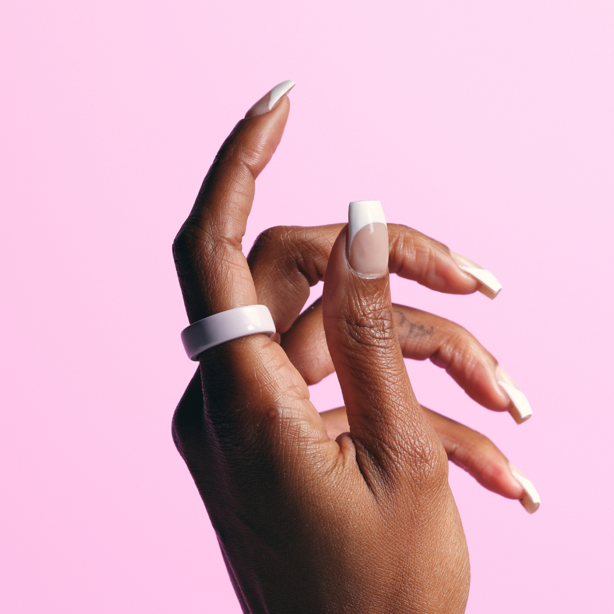 Eine Hand mit weiß bespitzten Nägeln und einem weißen Ring vor rosa Hintergrund.