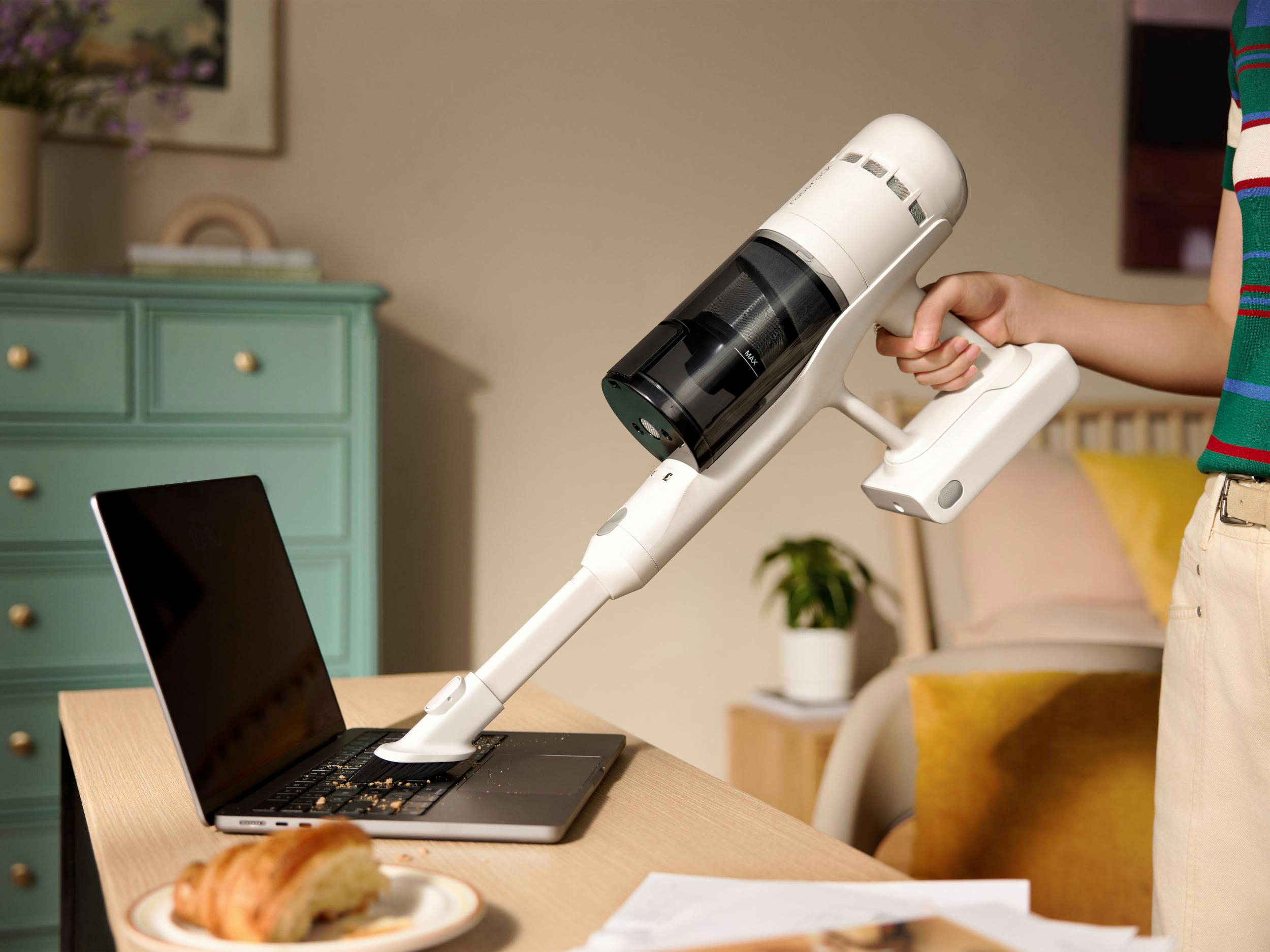 Eine Person saugt einen Laptop auf einem Schreibtisch. Ein Croissant liegt daneben. Im Hintergrund ist ein Minzschrank.