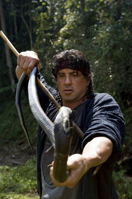 Sylvester Stallone hält eine Schlange mit einem Stock im Wald. Er trägt ein Stirnband und wirkt besorgt. Im Hintergrund sind Bäume zu sehen.
