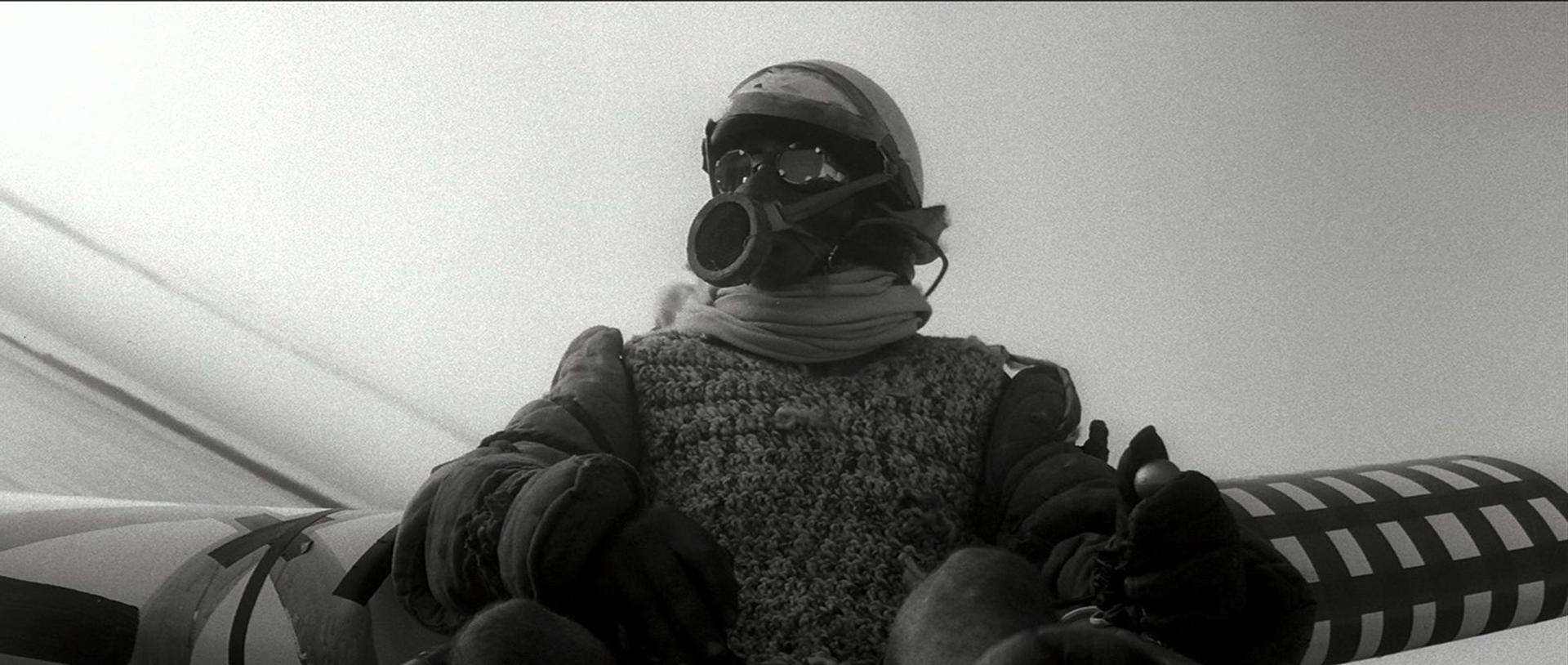 Eine Person mit Helm und Maske sitzt in einem Fahrzeug. Handschuhe und ein Strickpullover sind sichtbar. Der Hintergrund ist ein heller Himmel.
