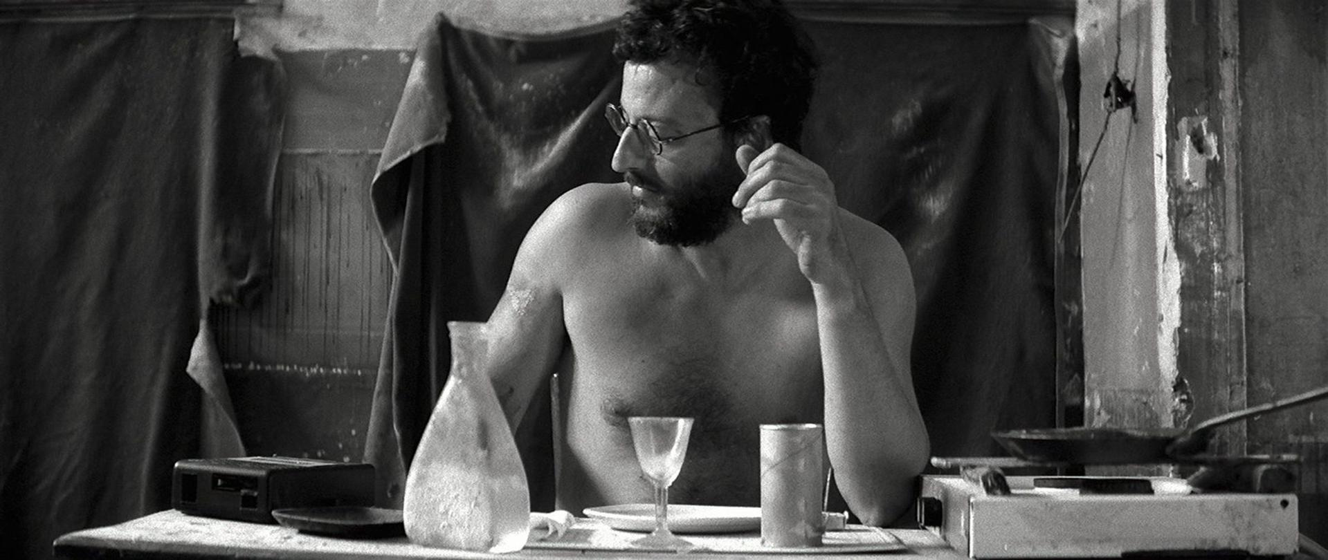 Ein Mann mit Brille und Bart sitzt an einem Tisch. Es gibt eine Kanne, ein Glas und einen Teller. Der Hintergrund ist dunkel und rustikal.