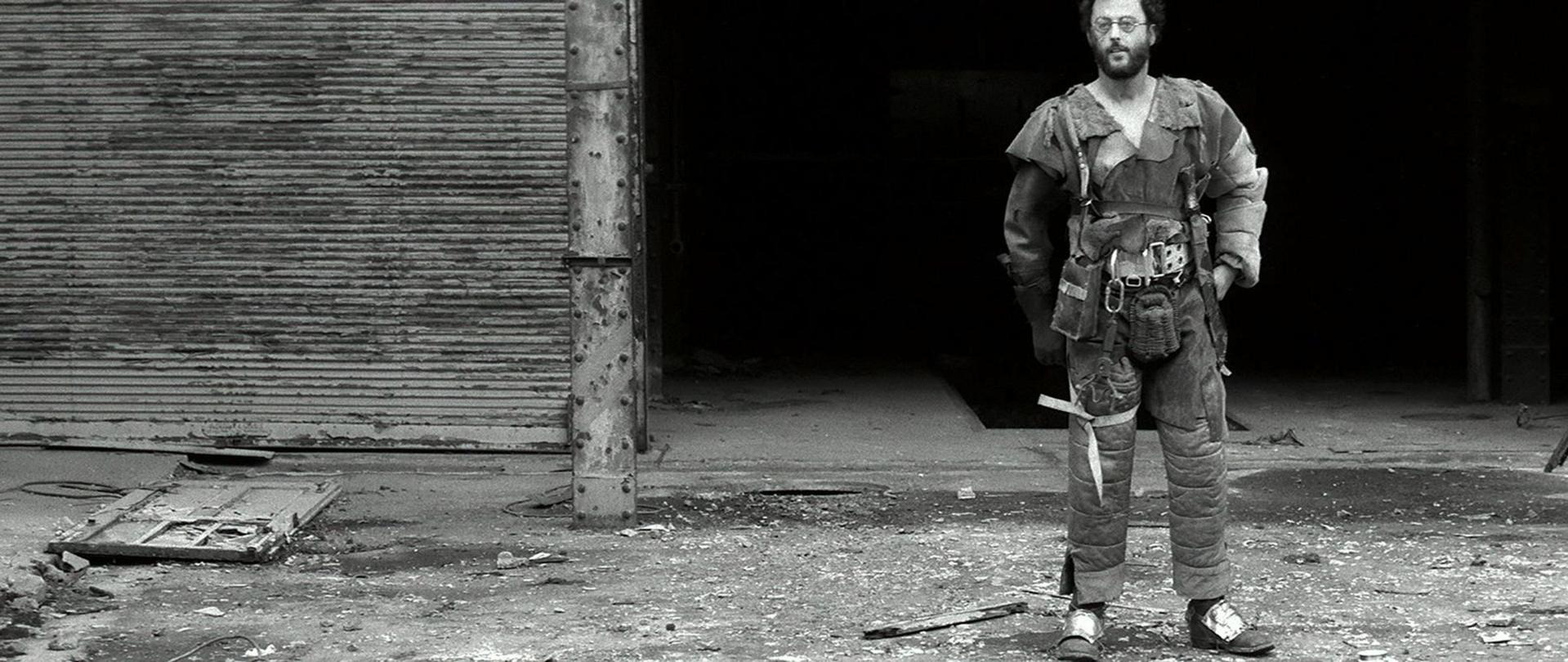 Ein Mann in abgenutzter Kleidung steht in einem verfallenden Gebäude. Eine zerbrochene Tür liegt am Boden. Der Hintergrund ist dunkel und schattig.