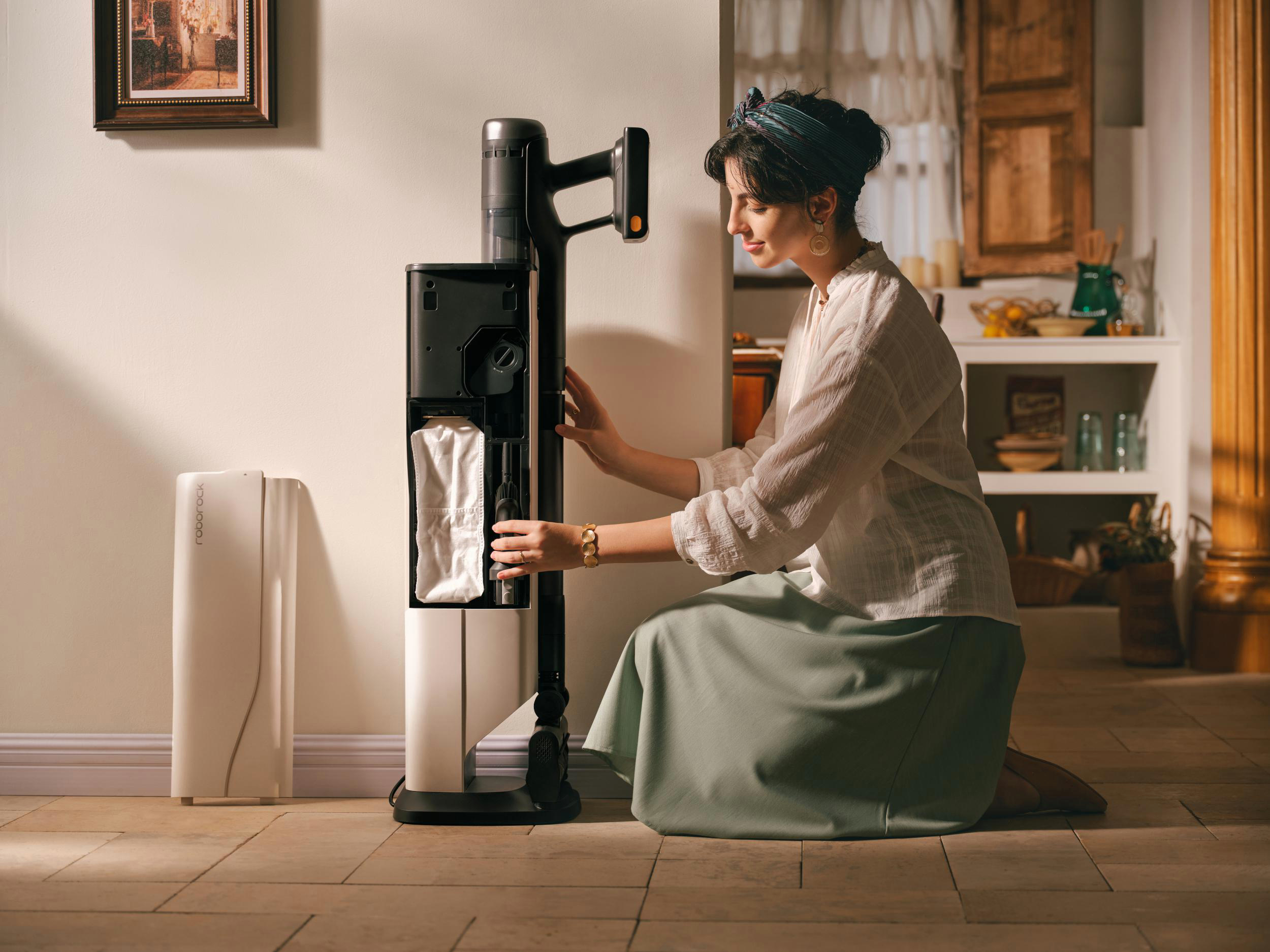 Eine Frau kniet und interagiert mit einem Staubsauger. Ein Gemälde hängt an der Wand. Der Hintergrund zeigt eine Küche.
