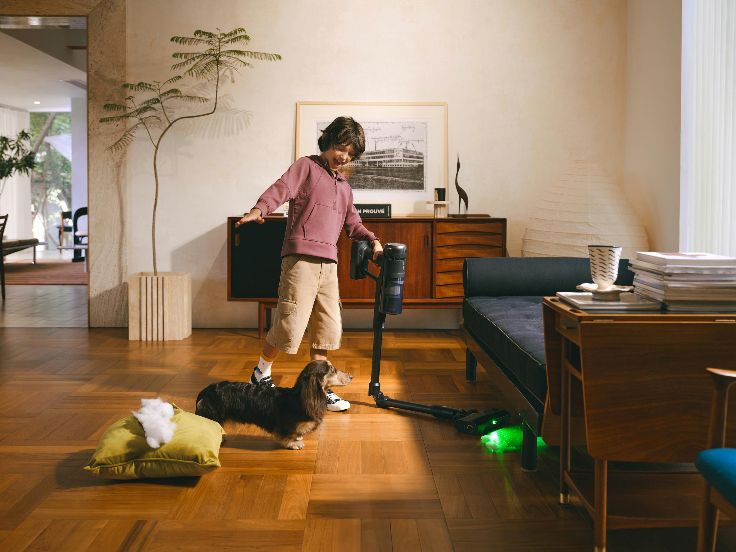 Ein Junge mit Staubsauger, Hund, gelbem Kissen und Möbeln in einem Raum.