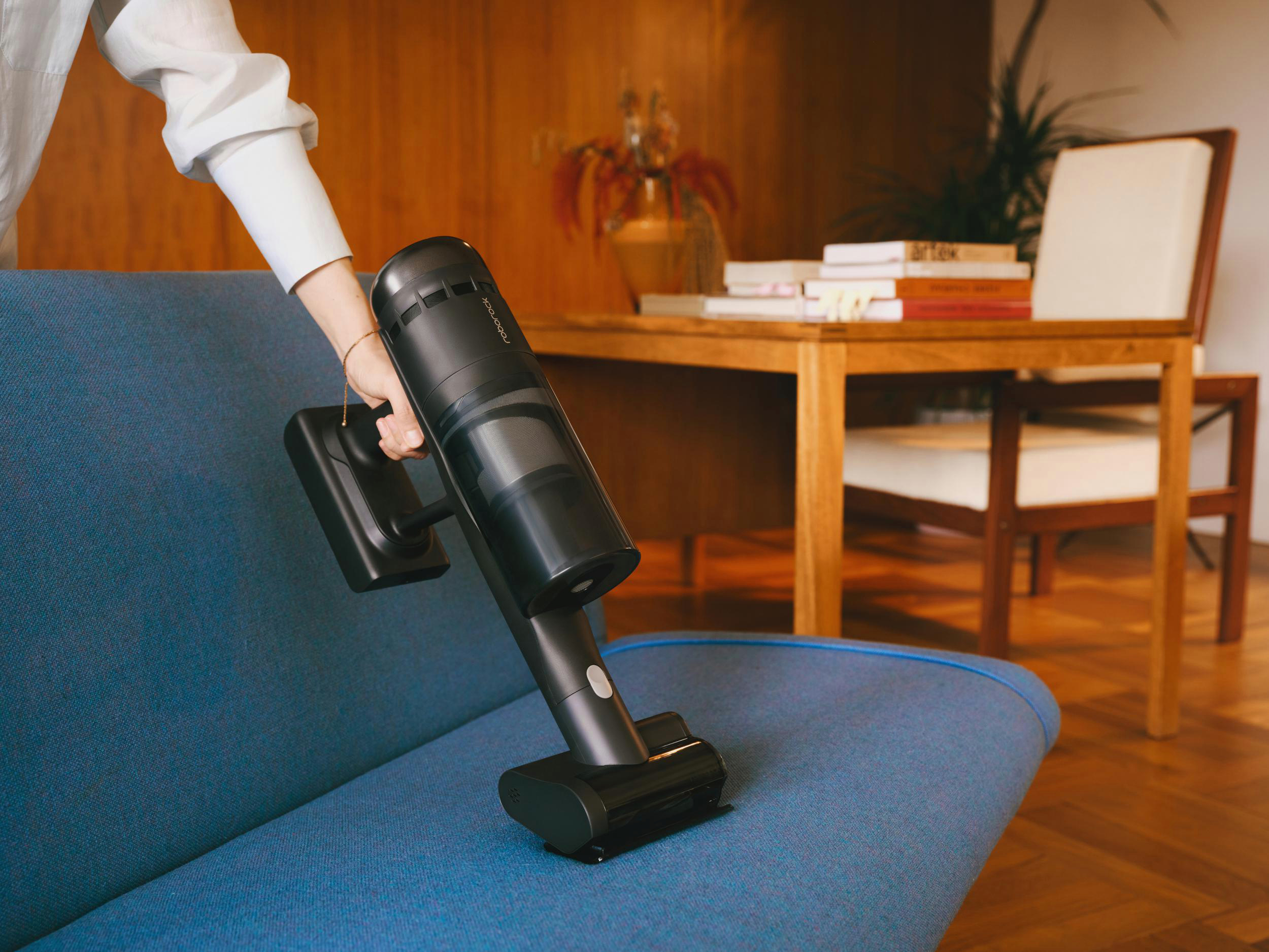 Eine Person saugt mit einem schwarzen Staubsauger ein blaues Sofa ab. Im Hintergrund sind ein Holztisch und ein Stuhl.