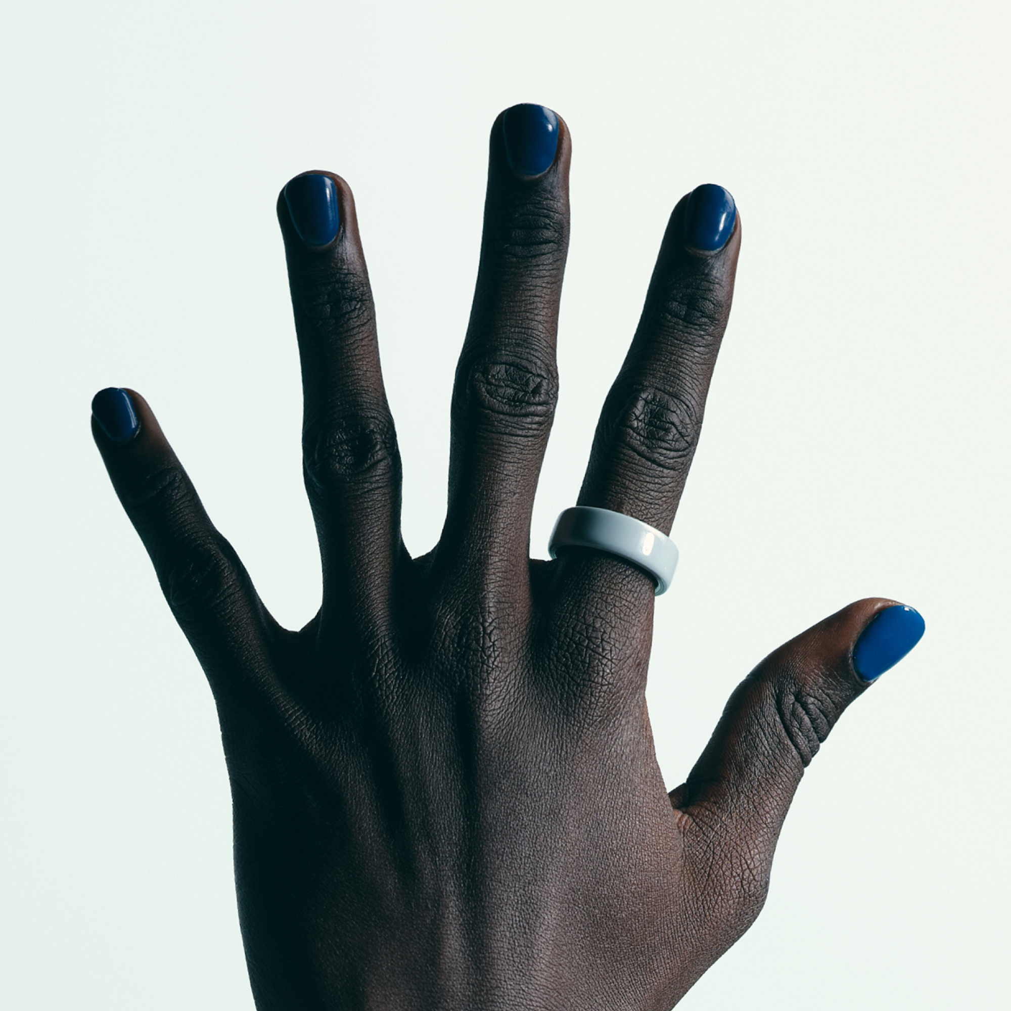 Eine dunkelhäutige Hand mit blauem Nagellack. Ein hellblauer Ring ziert den Mittelfinger. Der Hintergrund ist weiß.