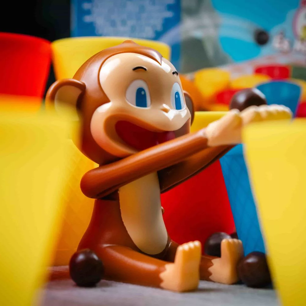 Egy Curious George figura színes csészék és szétszórt csokoládé golyók között ül. A majom mosolyog, kinyújtja a karjait.