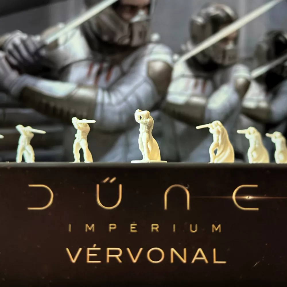 Miniatűr figurák a Dune: Imperium társasjátékból. Katonafigurák a játék címe előtt. A háttérben nagyobb kardos figurák.