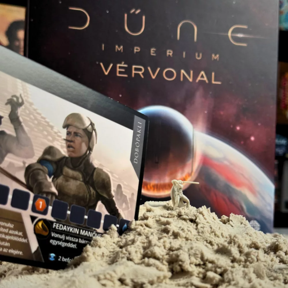 A Dune: Imperium társasjáték felállítása. A középpontban a Fedaykin Manőve szövegű kártya, homokon egy miniatúra, a háttérben a doboz.