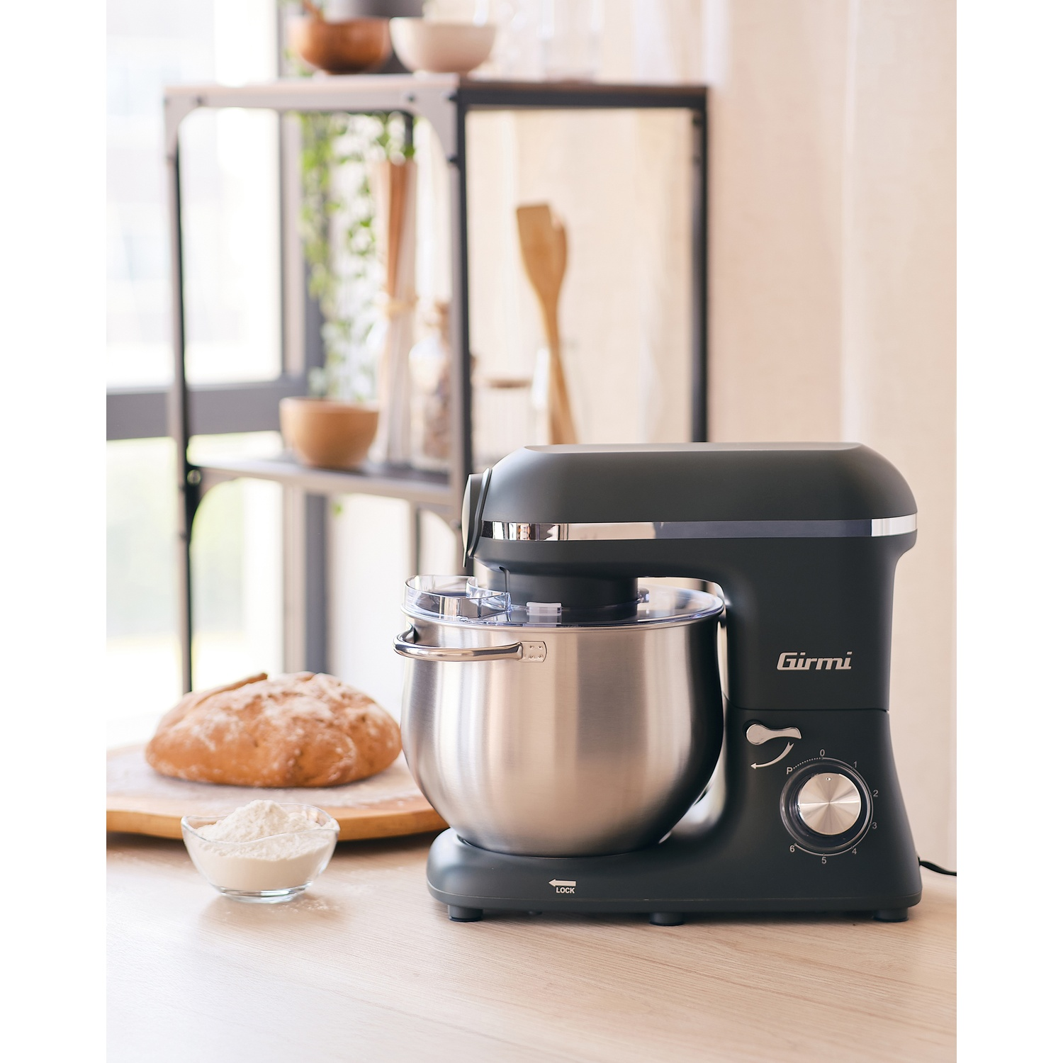 Un mixer nero su un tavolo di legno, vicino a una pagnotta e una ciotola di farina. Lo sfondo ha mensole con oggetti da cucina.