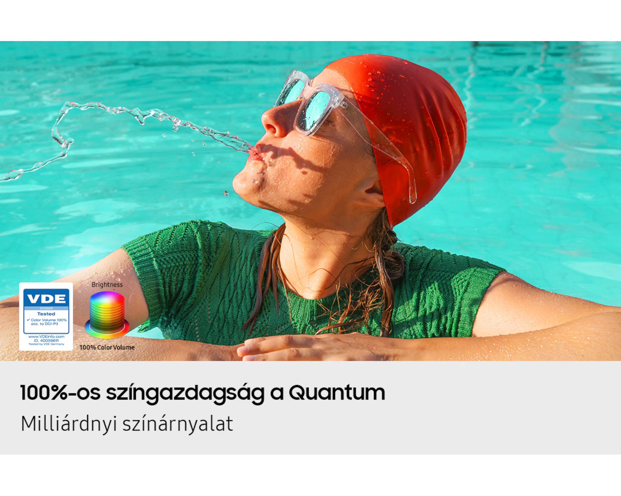 Egy személy medencében, piros úszósapkával és szemüveggel, vizet fúj ki a szájából. Zöld ing.