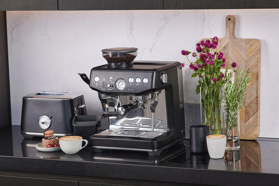 Eine schwarze Espressomaschine, ein Toaster und Kaffeetassen auf einer Arbeitsplatte. Blumen und ein Schneidebrett im Hintergrund.