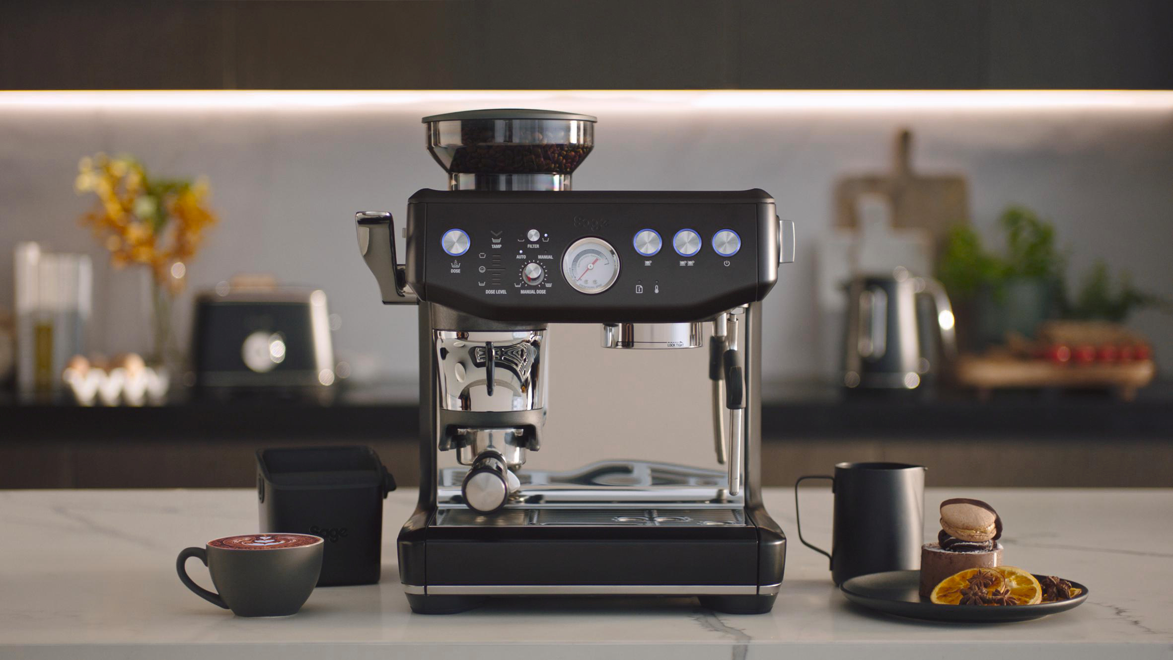 Eine schwarze Espressomaschine mit Kaffeebohnen, eine Tasse, Makronen und eine Tasse Kaffee-Kunst stehen auf einer Marmorarbeitsplatte.