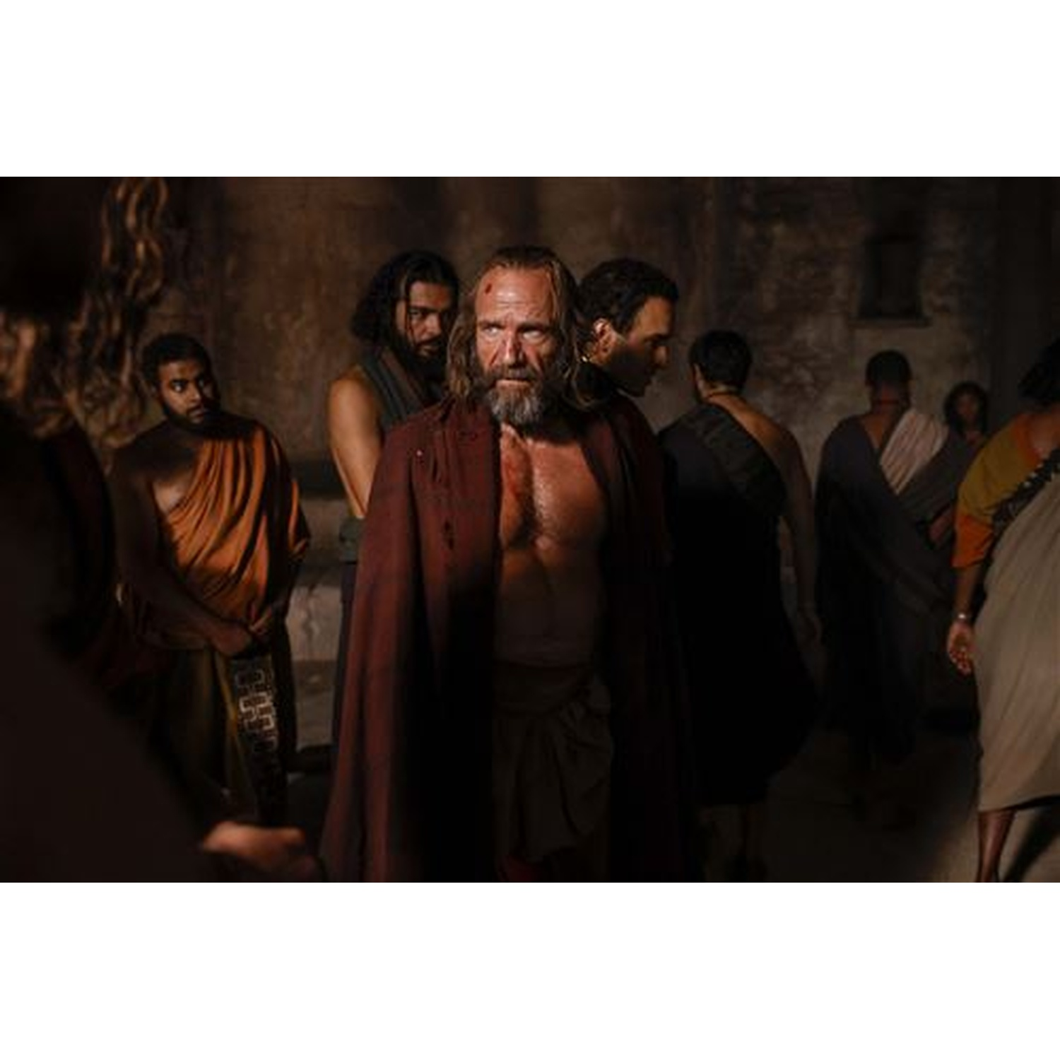 Un homme barbu avec un manteau rouge se tient parmi d'autres dans une pièce sombre. Certains portent des robes. La scène semble historique.
