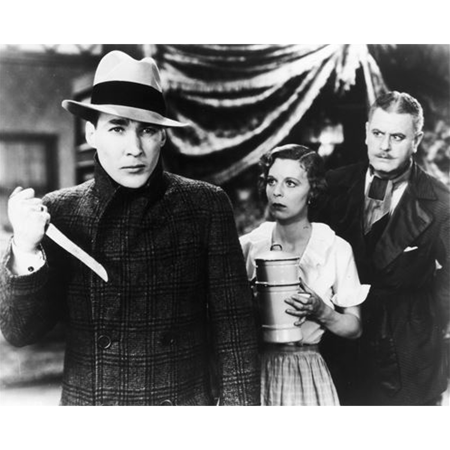 Un homme chapeauté tient un couteau, flanqué d'une femme avec un récipient et d'un autre homme. Scène de film en noir et blanc.