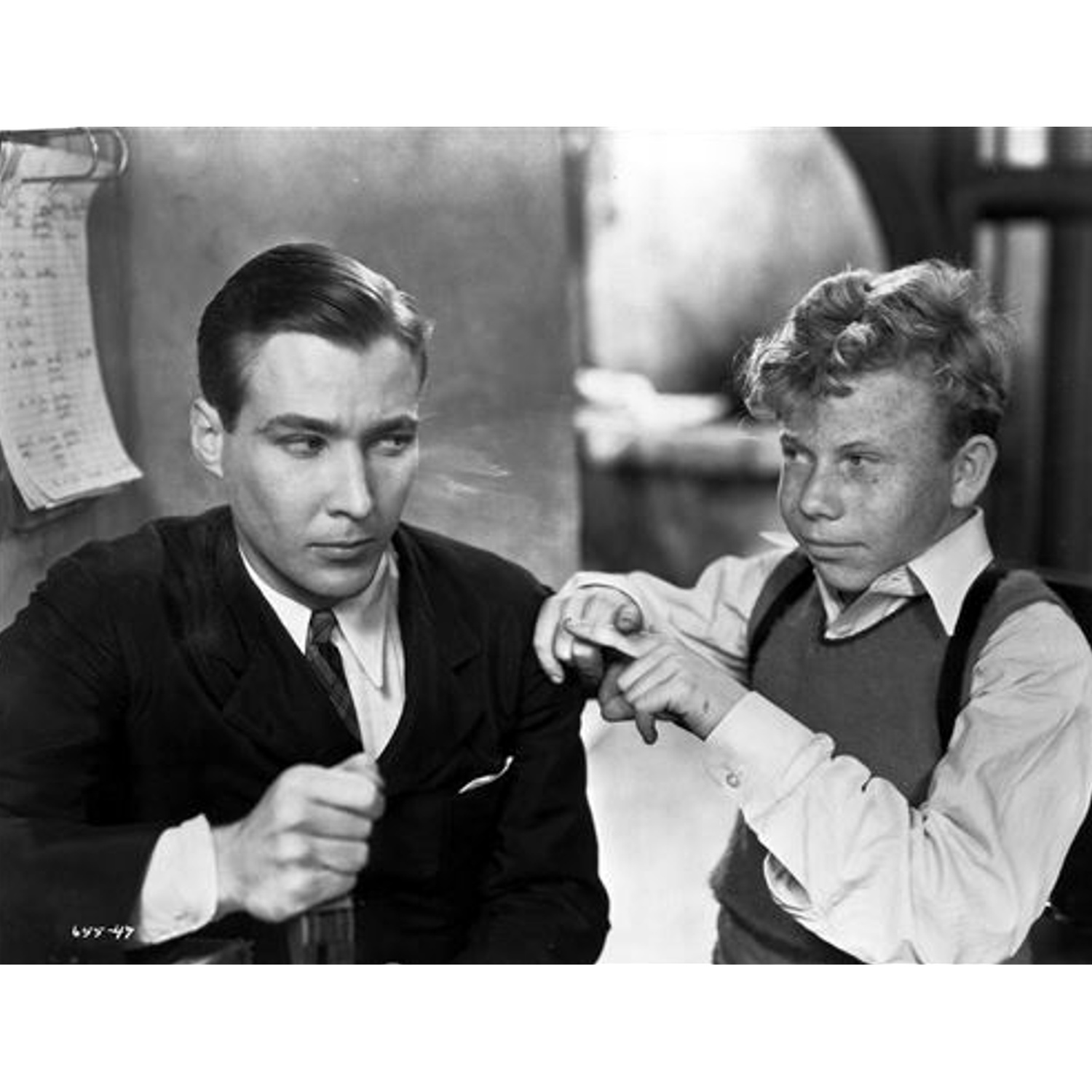 Deux hommes sur une photo en noir et blanc. L'un en costume et l'autre en gilet et bretelles. L'homme en costume tient une bouteille.