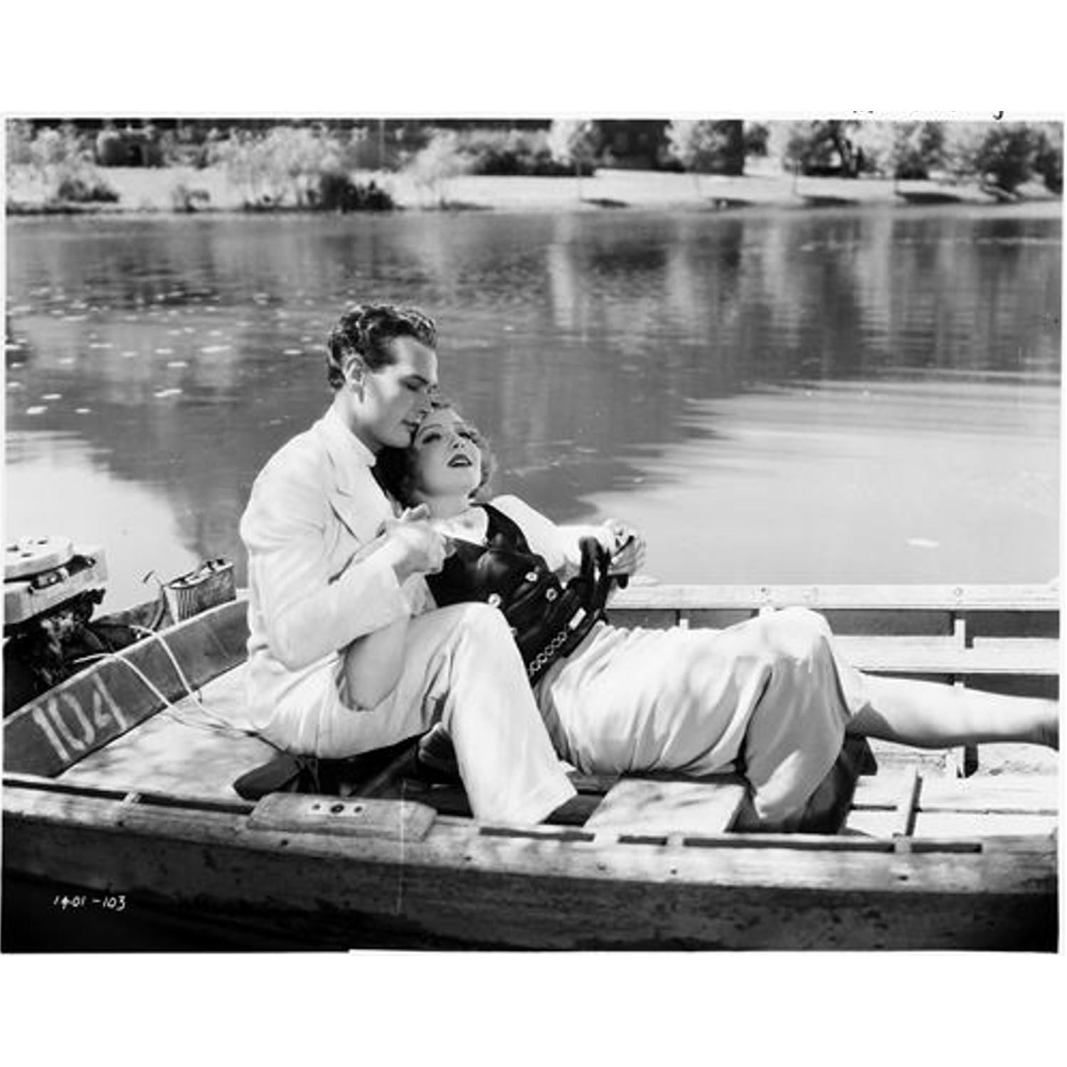 Un homme et une femme sont dans un bateau sur l'eau. L'homme en vêtements blancs embrasse la femme. Des bateaux et des arbres sont en arrière-plan.