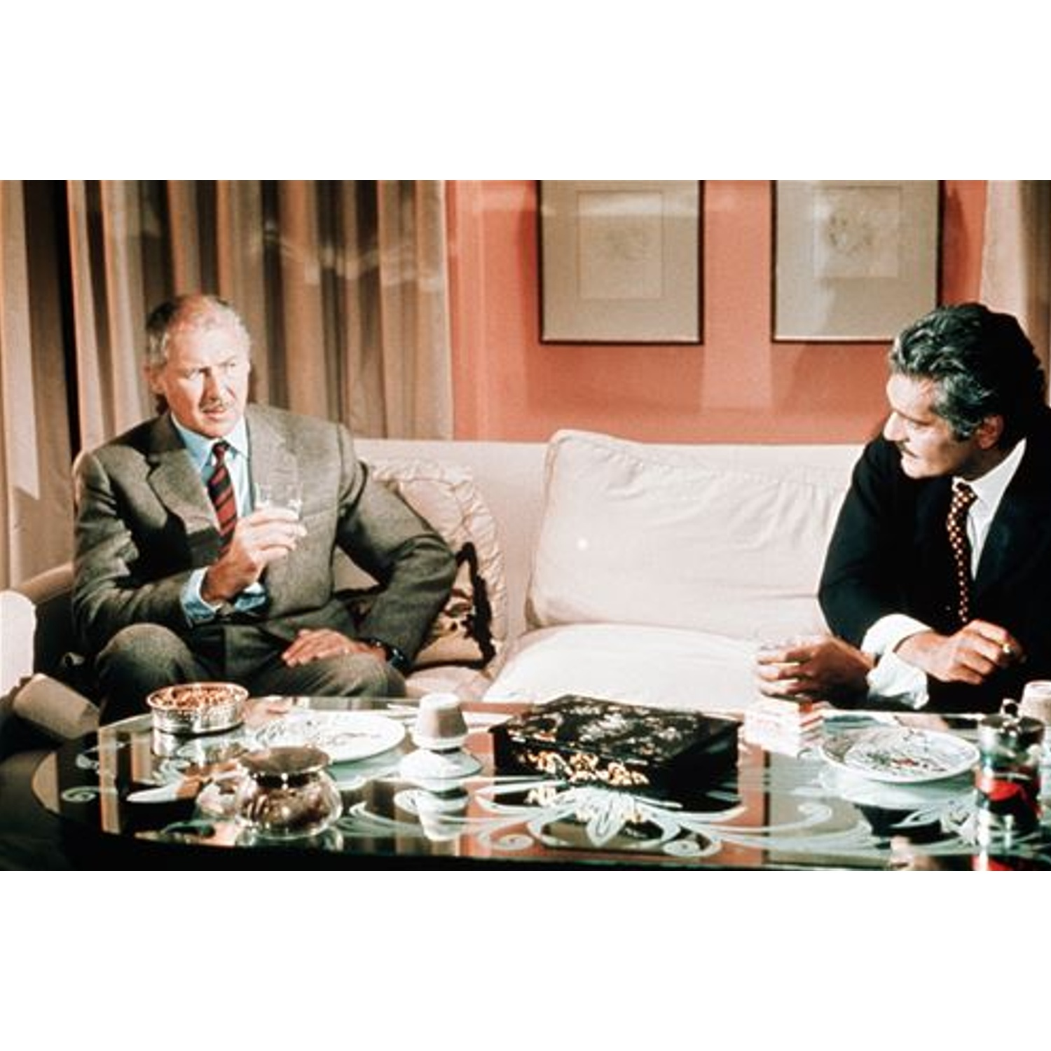 Deux hommes en costume sont assis sur un canapé, tenant des verres. Une table en verre devant a des plats et une boîte décorative. Fond avec art encadré.