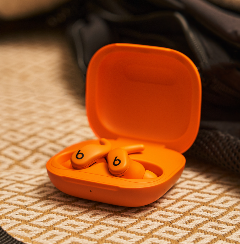 Orangefarbene Beats-Ohrhörer in einem offenen Etui. Das Etui und die Ohrhörer liegen auf einer gemusterten, beigefarbenen Oberfläche. Im Hintergrund ist ein dunkler Rucksack zu sehen.