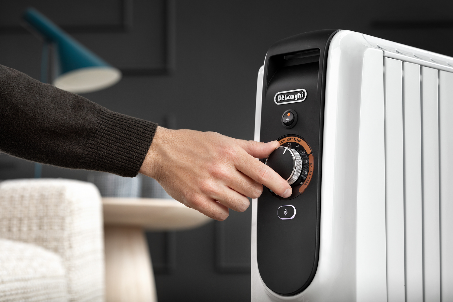Una mano gira el dial de un radiador de aceite De'Longhi blanco y negro. El dial indica los ajustes de temperatura. El fondo muestra una lámpara y un sofá.