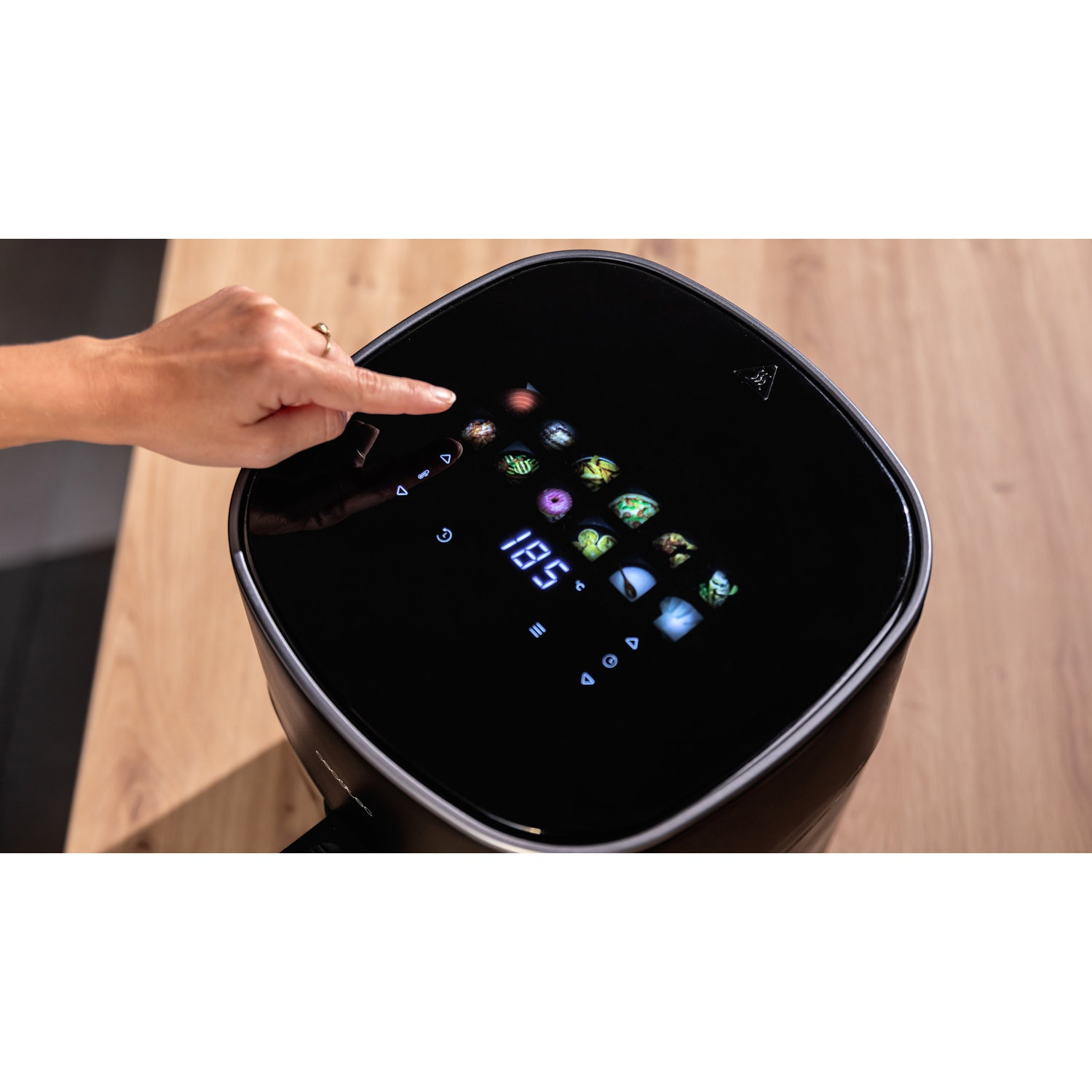Una mano tocca il touchscreen di una friggitrice ad aria nera. Lo schermo mostra le opzioni di cottura e la temperatura. Superficie in legno sullo sfondo.