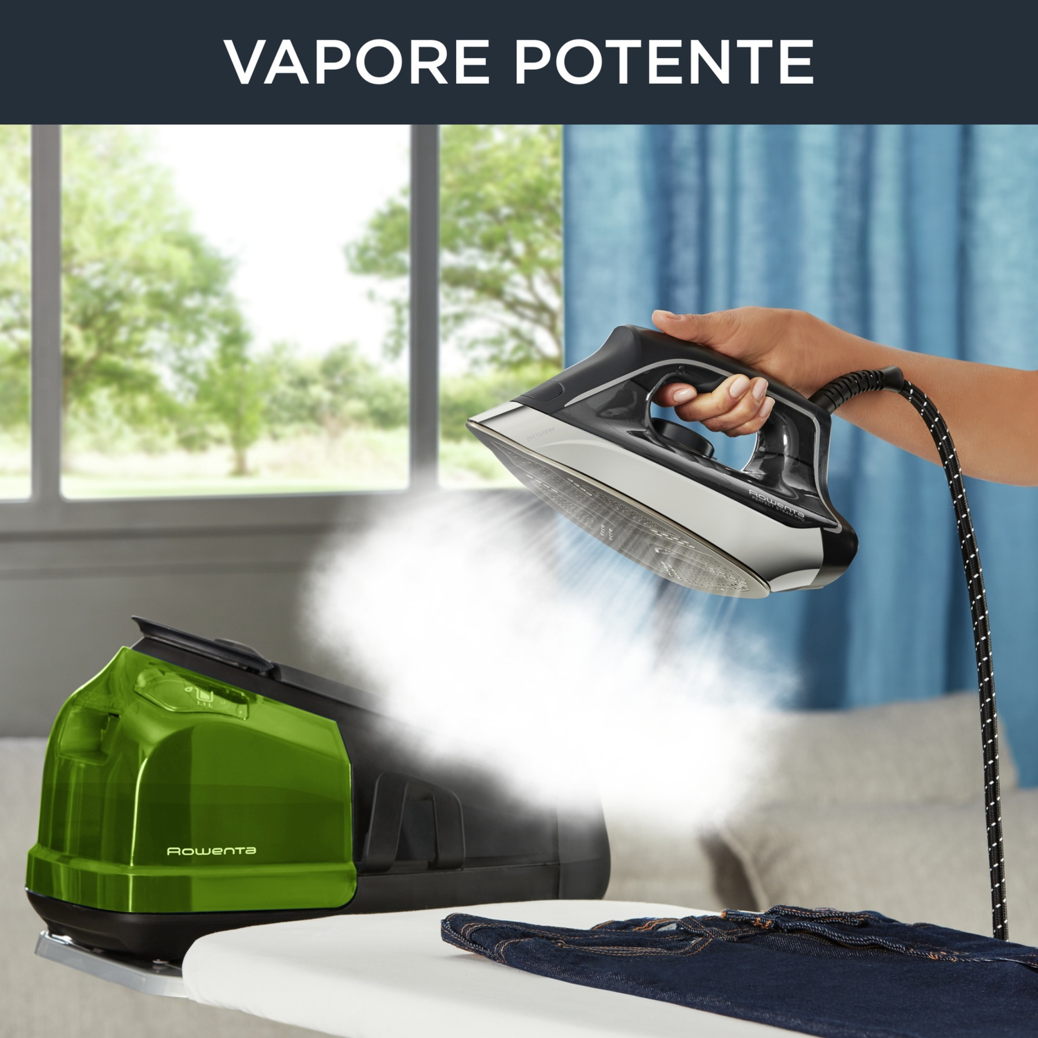 Una persona tiene in mano un ferro da stiro nero, esce vapore. Un generatore di vapore verde è su una tavola. Jeans sulla tavola.