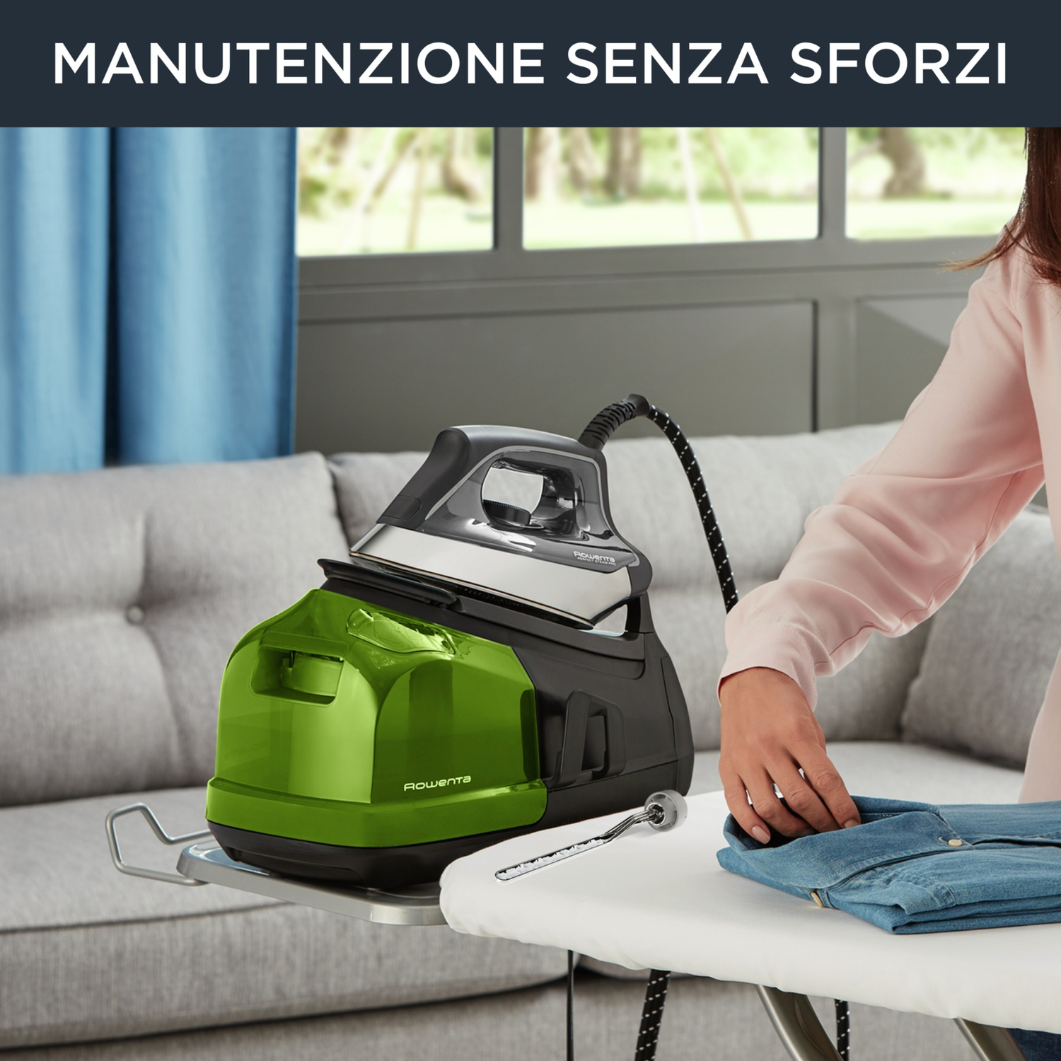 Un ferro da stiro con base verde su un asse da stiro. Una persona sta stirando una camicia blu. Lo sfondo ha una finestra e un divano.